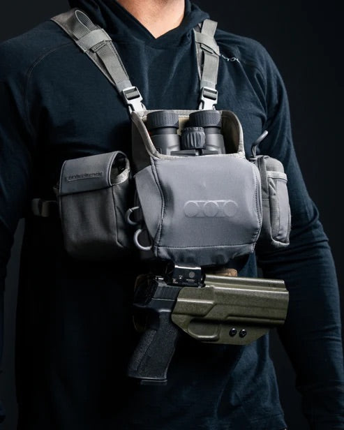 Eine Person mit schwarzem Kapuzenpulli modelliert das EBERLESTOCK Recon Modular Bino Pack Large mit Fernglas, Taschen und einer im Holster steckenden Handfeuerwaffe - und hebt es als unverzichtbare Jagdausrüstung vor einem dunklen Hintergrund hervor.