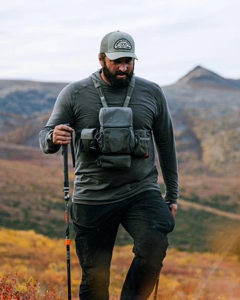 Ein Mann in Outdoor-Ausrüstung mit Wanderstock, Mütze und Rucksack mit dem EBERLESTOCK Recon Modular Bino Pack Large wandert durch eine malerische Berglandschaft mit herbstlich gefärbter Vegetation.