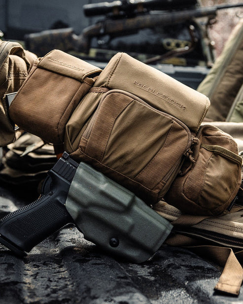 Das EBERLESTOCK Recon Modular Bino Pack Large in taktischem Hellbraun ruht auf einer Jagdausrüstung, mit einer schwarzen Handfeuerwaffe in einem grünen Holster im Vordergrund und einem Gewehr mit Zielfernrohr im Hintergrund.