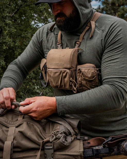 Ein bärtiger Mann in grünem Kapuzenhemd und Mütze passt sein EBERLESTOCK Recon Modular Bino Pack Large an, während er einen großen olivgrünen Rucksack in der freien Natur, umgeben von Bäumen, handhabt - ideal, um die gesamte Jagdausrüstung zu transportieren.