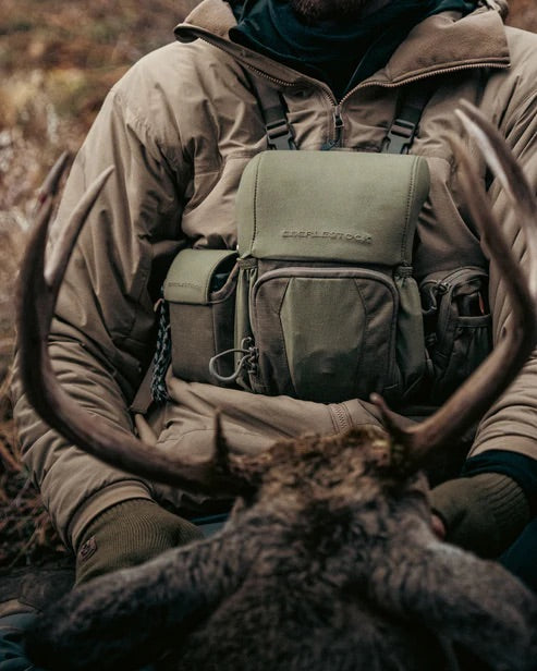 Eine Person in Outdoor-Ausrüstung hält ein Hirschgeweih mit einem grünen Jagdrucksack, Jagdausrüstung und einem EBERLESTOCK Recon Modular Bino Pack Large auf der Brust in einer natürlichen Umgebung.