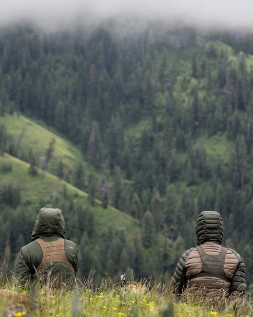 Zwei Personen in Kapuzenjacken, ausgestattet mit dem EBERLESTOCK Recon Modular Bino Pack Large, sitzen im hohen Gras und blicken auf einen nebligen Waldhügel. Der wolkenverhangene Himmel über ihnen hebt ihre Jagdausrüstung und die bewährte EBERLESTOCK Ausrüstung hervor.