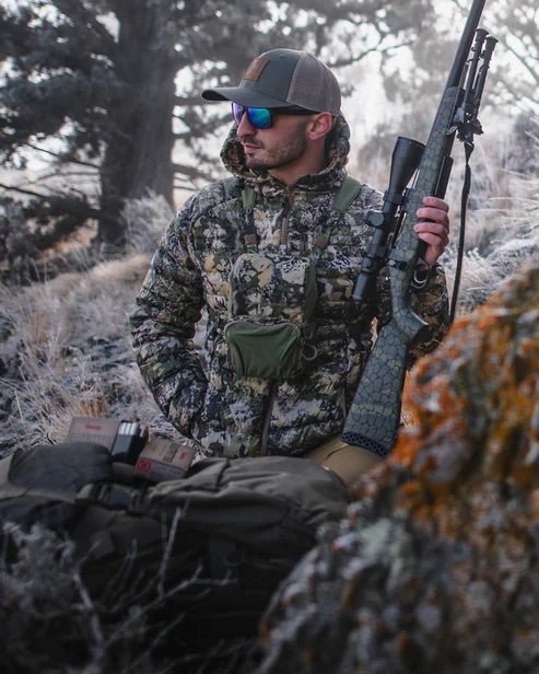 Ein Mann in Tarnkleidung und mit Sonnenbrille steht mit einem Gewehr im Freien, trägt einen Hut, eine grüne Brusttasche und das EBERLESTOCK Recon Modular Bino Pack Large inmitten seiner Jagdausrüstung in einer frostigen Graslandschaft.