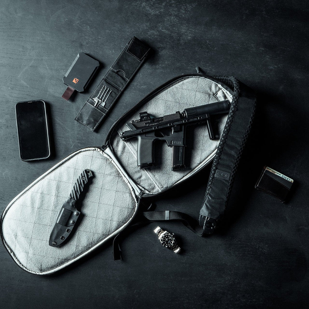 Draufsicht auf einen offenen EBERLESTOCK Fade Sling mit gesteppter grauer Innenseite, der eine Flux-Pistole und ein Messer zum diskreten Tragen enthält, umgeben von einem Smartphone, einer Uhr, einer Brieftasche, einem Kartenhalter und einer Werkzeugrolle auf einer dunklen Oberfläche.