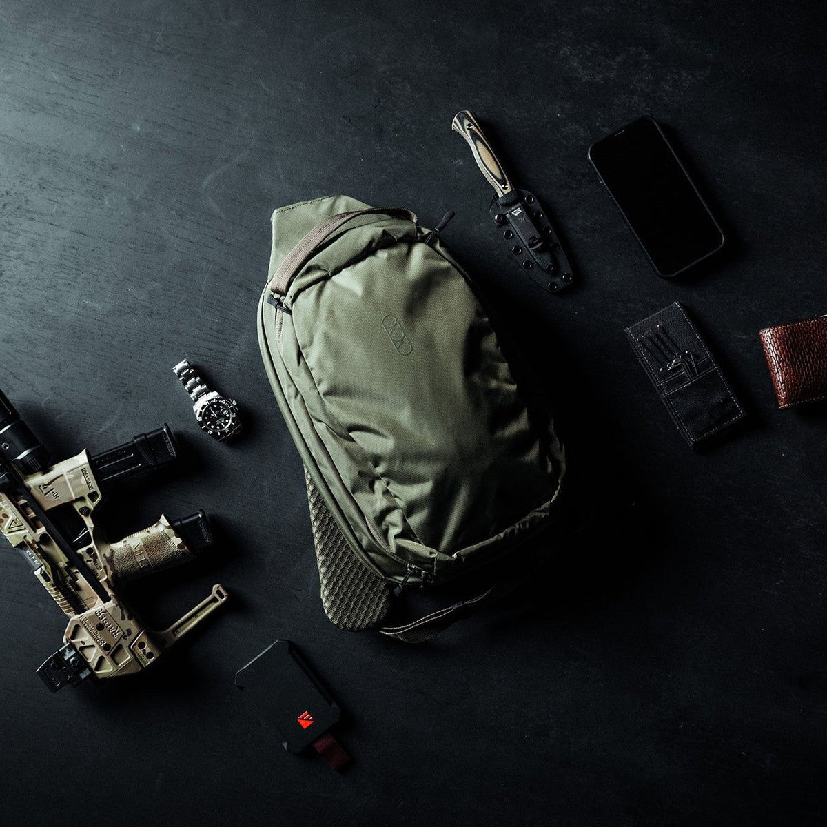 Ein olivgrüner EBERLESTOCK Fade Sling Rucksack, ein taktisches Messer, ein schwarzes Smartphone, zwei Brieftaschen, eine Uhr, ein Akkupack und ein hellbraunes Airsoft-Gewehr sind auf einer dunklen Fläche angeordnet.