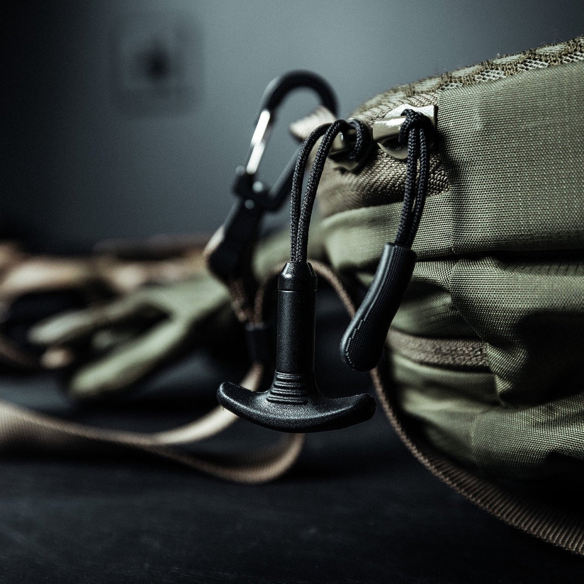 Nahaufnahme eines grün-braunen Rucksacks mit dem EBERLESTOCK Eberlestock Fade Sling und einem am Gurt befestigten Karabiner, sowie einem schwarzen Reißverschluss. Die Tasche steht auf einer dunklen Fläche mit einem leicht verschwommenen Hintergrund.