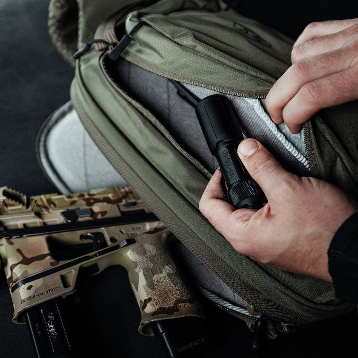 Eine Person steckt eine Taschenlampe in die Vordertasche eines geöffneten grünen EBERLESTOCK Fade Sling, unter dem eine braun getarnte Handfeuerwaffe und ein schwarzes Magazin teilweise sichtbar sind - ideal zum diskretes Pistolen Tragen.