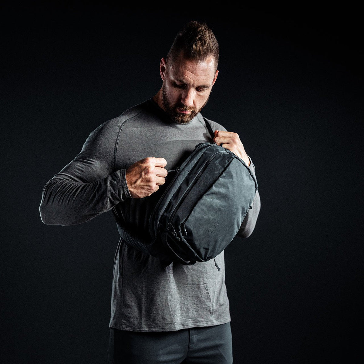 Ein Mann in einem taillierten grauen langärmeligen Hemd stellt vor einem dunklen Hintergrund den Riemen eines EBERLESTOCK Fade Sling ein, der sich ideal zum diskreten Tragen einer Pistole eignet.