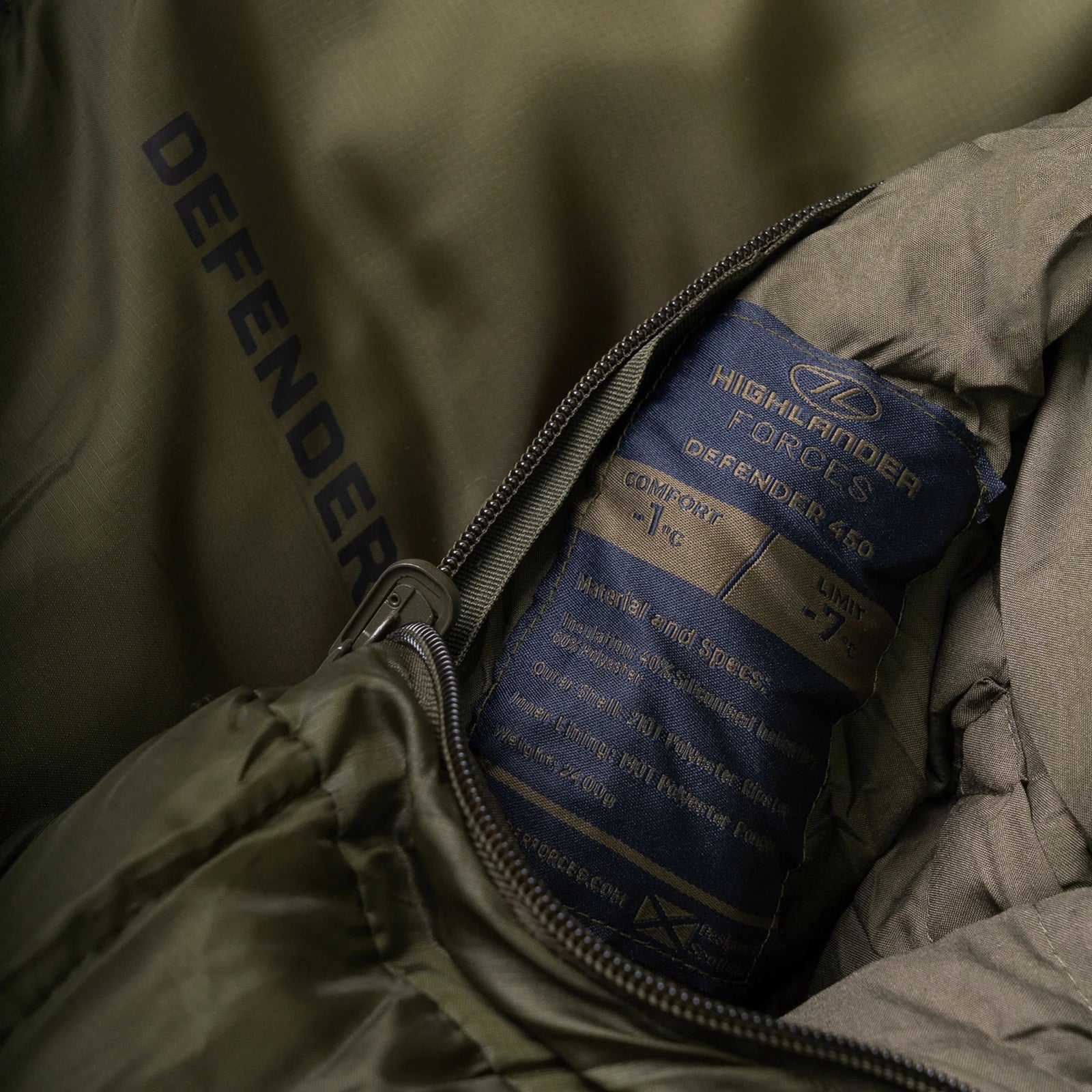 Nahaufnahme eines grünen Highlander Schlafsacks Defender 450 von HIGHLANDER OUTDOOR, Etikett mit Angaben zu Komfort und Grenztemperaturen, mit "DEFENDER"-Aufdruck innen neben dem Reißverschluss. Ideal als 4-Jahreszeiten-Schlafsack.