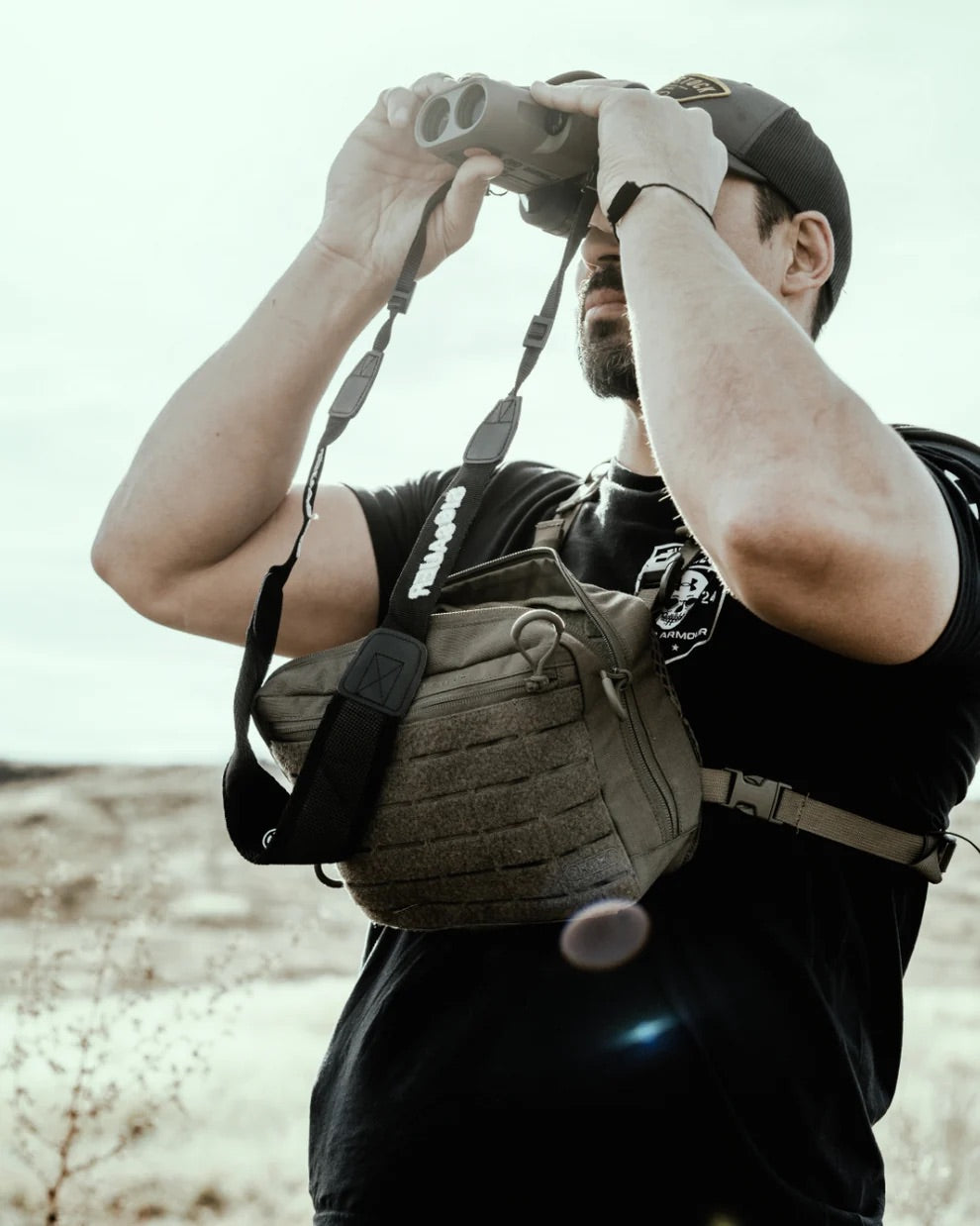 Eine Person, die ein schwarzes Hemd, eine Mütze und den EBERLESTOCK Force Chest Pack trägt, schaut durch ein Fernglas in eine trockene, sonnige Landschaft.