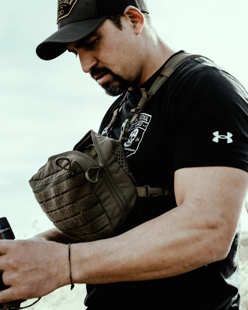 Ein Mann im Freien, der eine schwarze Mütze, ein schwarzes Under Armour T-Shirt und eine taktische Tragetasche trägt, justiert sein EBERLESTOCK Force Chest Pack.