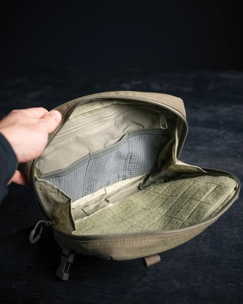 Eine Hand hält einen leeren olivgrünen EBERLESTOCK Force Chest Pack auf, der vor einem dunklen Hintergrund seine organisierten, MOLLE-kompatiblen Netz- und Stofftaschen im Inneren offenbart.