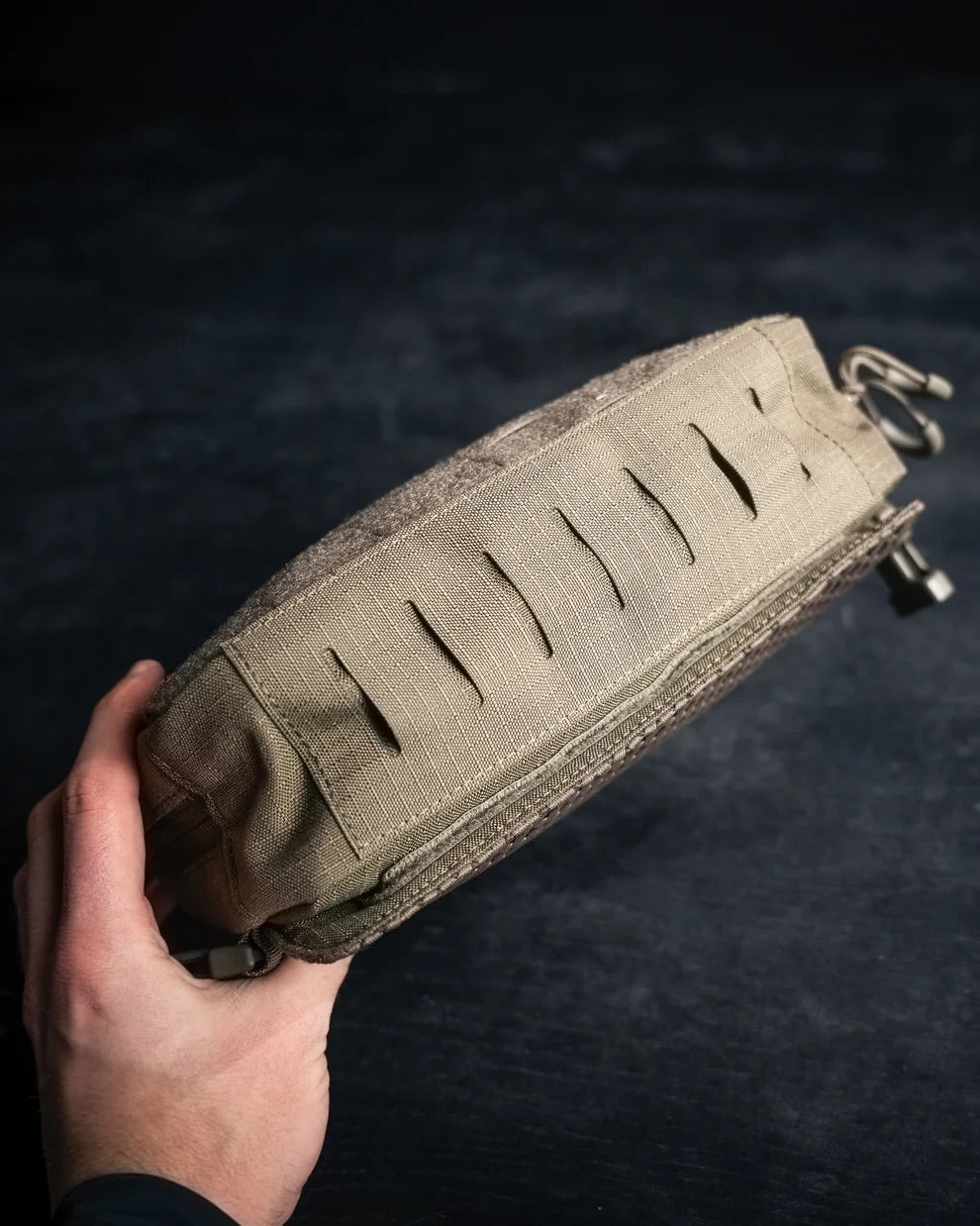 Eine Person hält ein EBERLESTOCK Force Chest Pack, eine hellbraune taktische Tasche mit horizontalen Schlitzen für modulare Befestigung und MOLLE-Kompatibilität, vor einem dunklen, unscharfen Hintergrund.