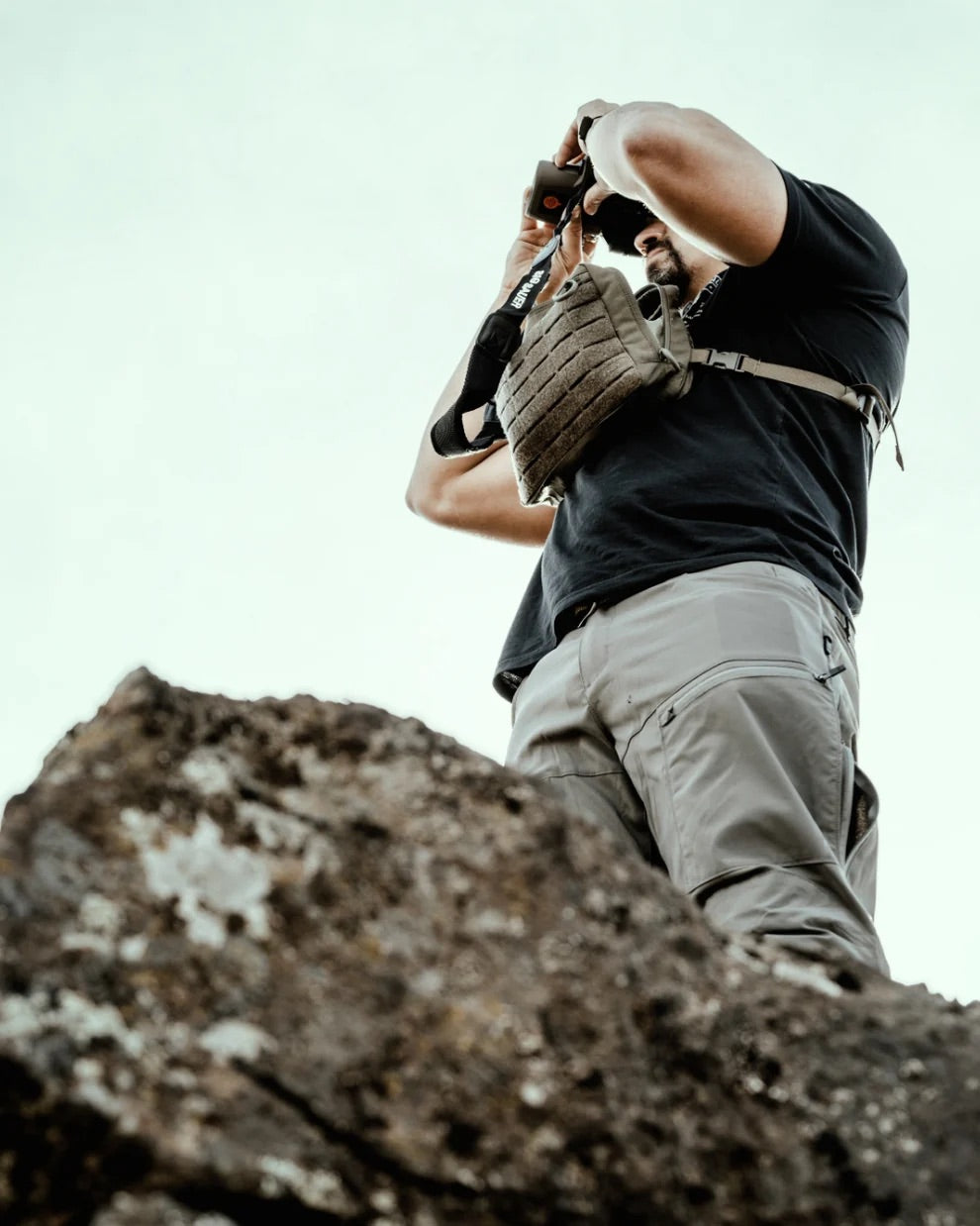 Eine Person in Outdoor-Kleidung steht auf einer felsigen Fläche und blickt durch ein Fernglas nach oben. Sie trägt ein schwarzes Hemd, eine helle Cargohose und den EBERLESTOCK Force Chest Pack - ein MOLLE-kompatibler Chest Pack für die Jagd. Hinter ihnen ist der Himmel klar.
