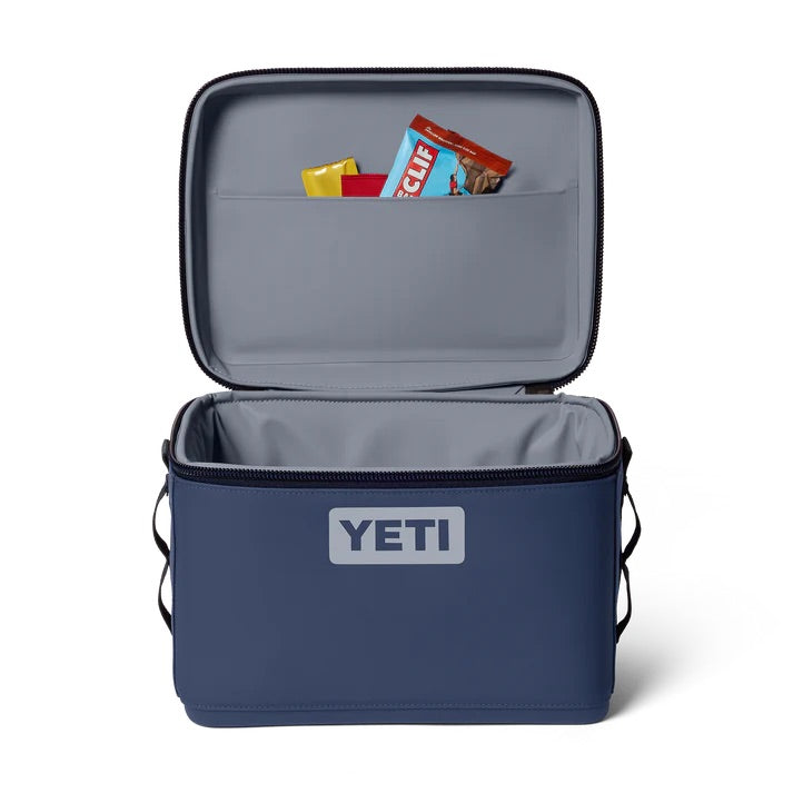 Eine YETI® Kühltasche Daytrip 9L in Classic Navy liegt offen da und zeigt zwei Müsliriegel und ein gelbes Päckchen im oberen Fach und ein leeres Hauptfach darunter.