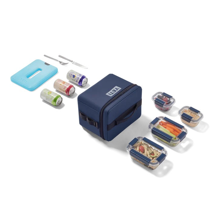 Die YETI® Kühltasche Daytrip 9L - Classic Navy von YETI wird mit drei rechteckigen Lebensmittelbehältern aus Glas, verschiedenen Lebensmitteln, drei Getränkedosen, einem blauen Eisbeutel und Utensilien auf weißem Hintergrund dargestellt.