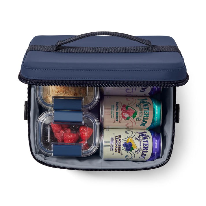 Die YETI® Daytrip 9L Kühltasche in Classic Navy mit Reißverschluss bietet Platz für zwei Dosen Sprudelwasser, einen durchsichtigen Behälter mit Himbeeren und Brombeeren sowie zwei durchsichtige Behälter mit Müsli, die alle übersichtlich angeordnet sind.