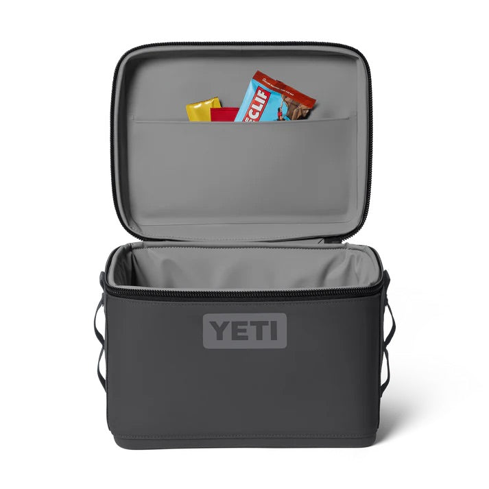 Eine anthrazitfarbene YETI® Kühltasche Daytrip 9L von YETI liegt offen da und zeigt ihr graues Innenleben; in der Deckeltasche sind Snack-Riegel und Gewürzpakete untergebracht, während das Hauptfach dieser isolierten Lunchbag leer ist.