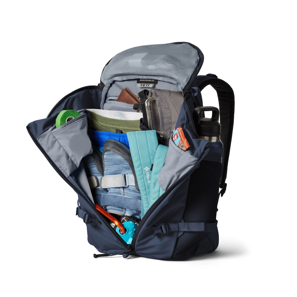 Ein YETI® Rucksack Ranchero™ 27L in Classic Navy wird weit geöffnet und gibt den Blick frei auf ordentlich verpackte Kleidung, eine Wasserflasche, Toilettenartikel, Schuhe, Sonnenbrille und Zahnpasta in Reisegröße in seinen organisierten Fächern.