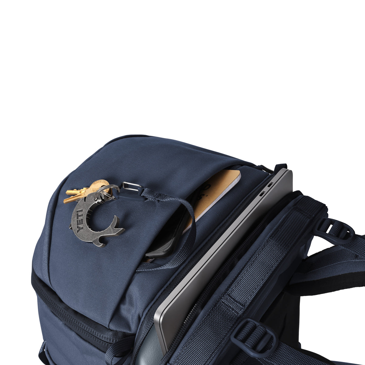 Ein YETI® Rucksack Ranchero™ 27L in Classic Navy mit geöffneter Deckeltasche, in der ein Smartphone und ein Tablet zu sehen sind, und einem Delphin-Schlüsselanhänger am Reißverschluss. Hergestellt aus TuffSkin Nylon für einen wasserfesten Transport.