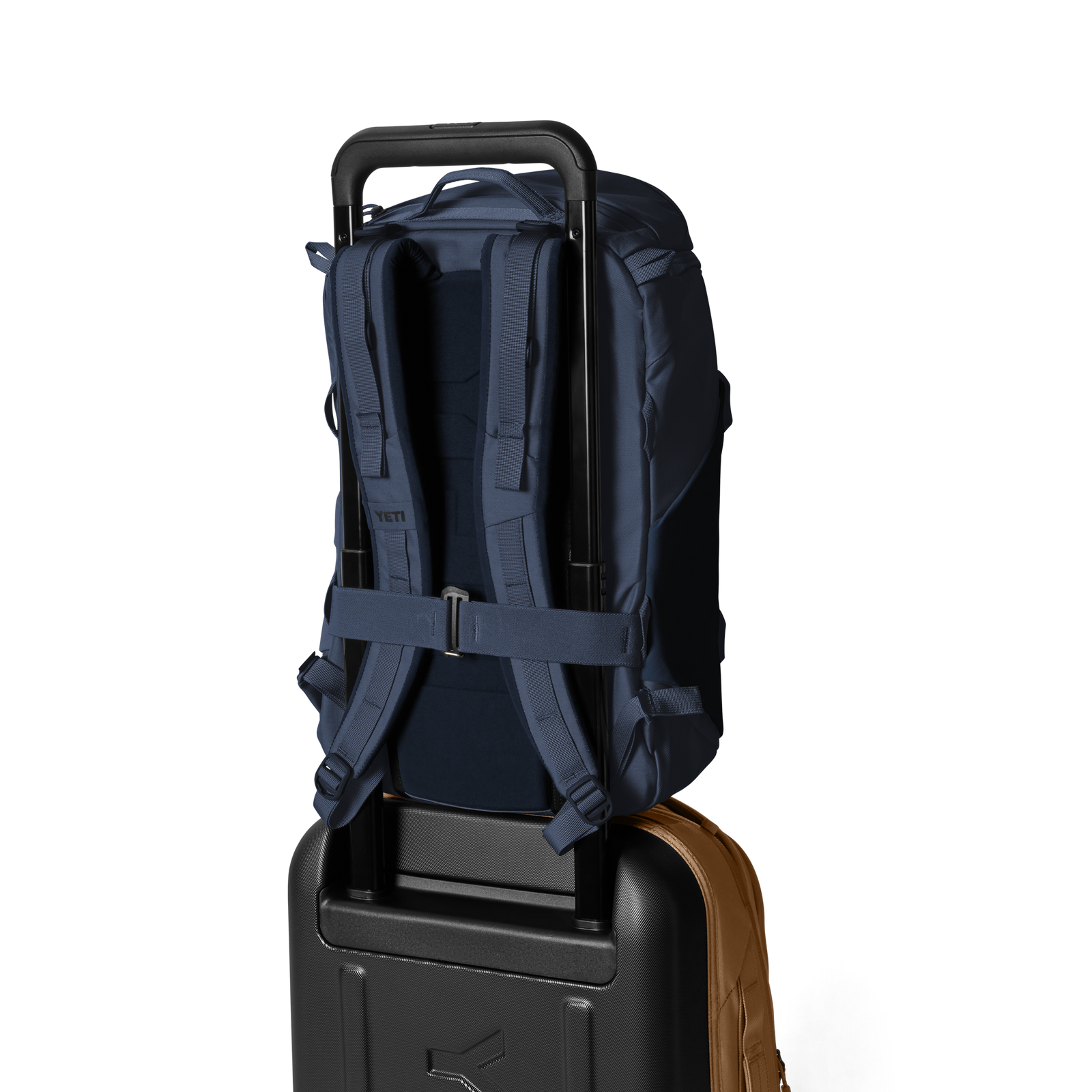 Der YETI® Rucksack Ranchero™ 22L in Classic Navy mit gepolsterten Schultergurten und einem Laptopfach sitzt sicher auf einem schwarzen Rollkoffer, zusammen mit einer braunen Tasche, alles vor einem schlichten weißen Hintergrund.