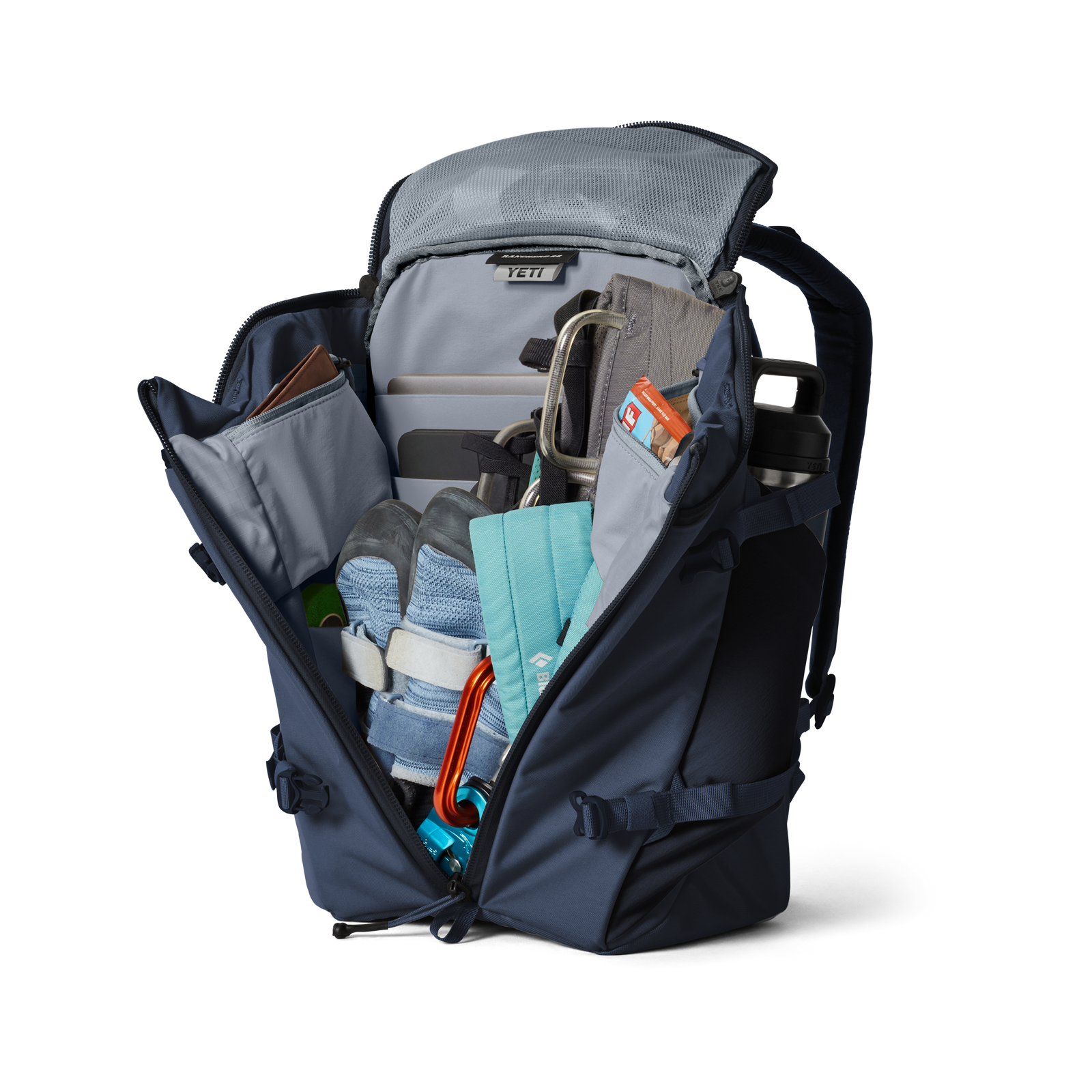 Ein YETI® Rucksack Ranchero™ 22L in Classic Navy wird vor einem schlichten weißen Hintergrund aufgeklappt und gibt den Blick auf den organisierten Inhalt frei: Schuhe, eine Wasserflasche, Bücher, eine Brieftasche, eine türkisfarbene Tasche, Gurte, Kletterkarabiner und ein Laptopfach.