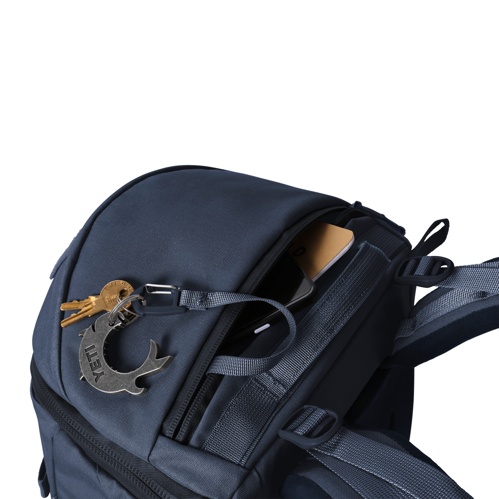 Der YETI® Rucksack Ranchero™ 22L in Classic Navy ist ein wasserdichter Rucksack mit einem Laptopfach, einer kleinen Reißverschlusstasche mit sichtbarem Telefon und Portemonnaie sowie einem Schlüsselanhänger mit Schlüsseln und Metallwerkzeug am Reißverschluss. Der Hintergrund ist weiß.