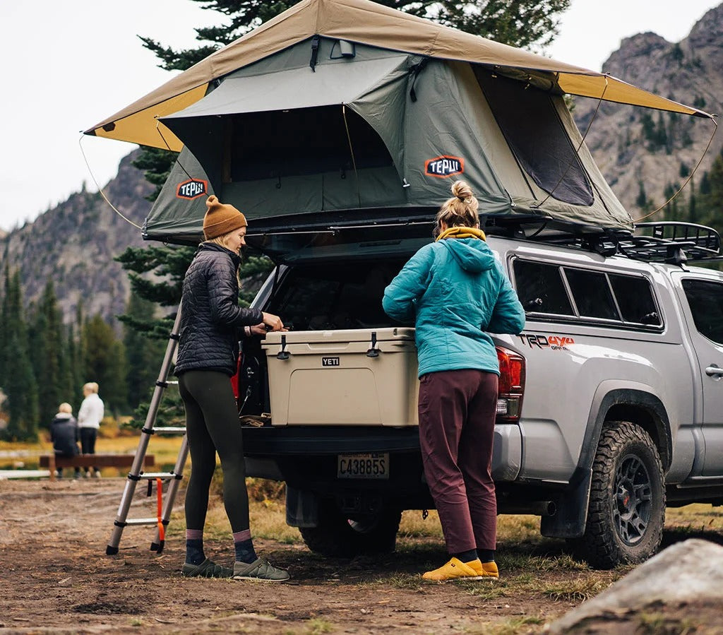 Zwei Personen stehen neben einem grauen Pick-up mit einem Dachzelt und laden eine große, hellbraune YETI®-Kühlbox „Tundra 75“ auf einem von Bäumen und Bergen umgebenen Campingplatz aus. Campingausrüstung ist zu sehen, und es ist ein bewölkter Tag.