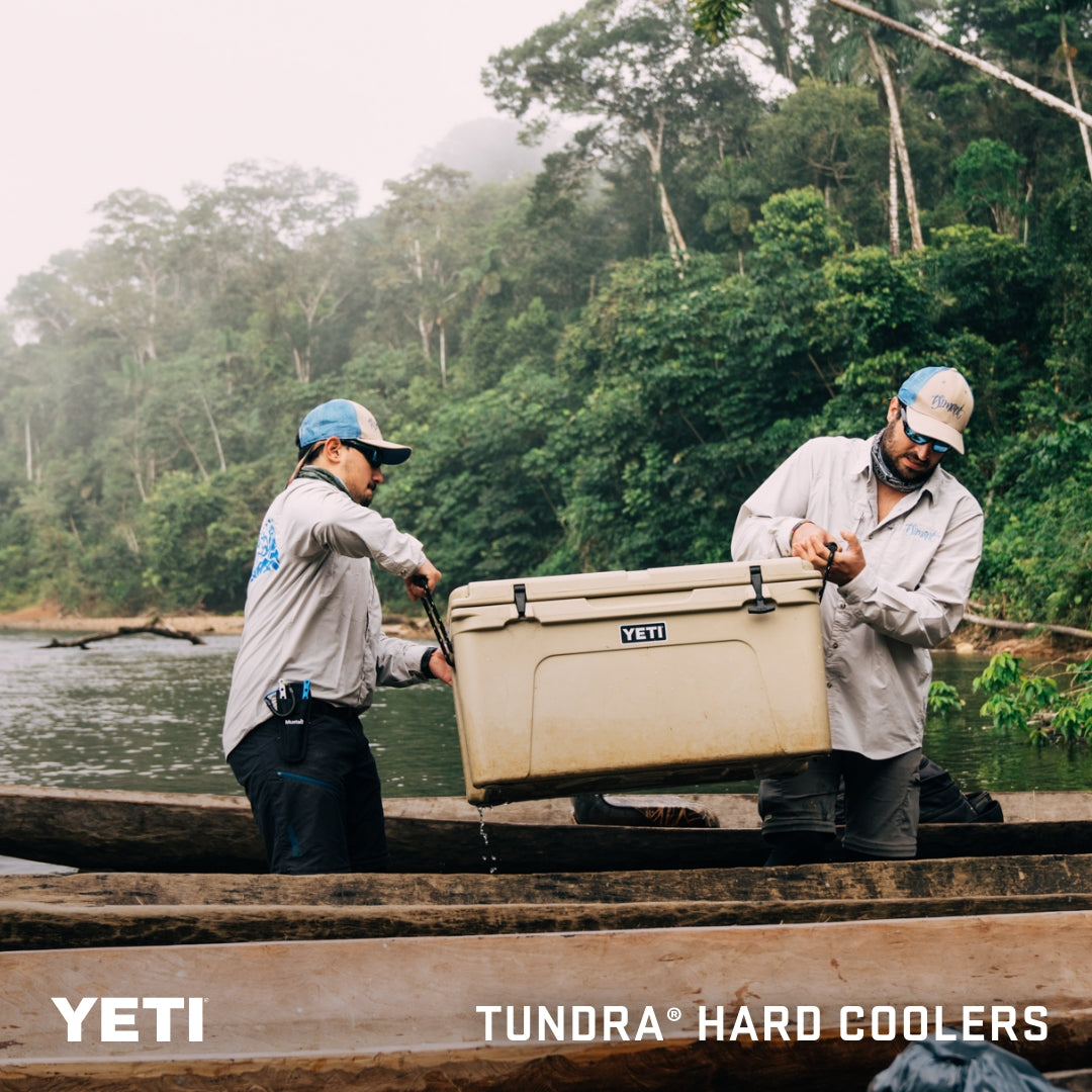 Zwei Personen in Outdoor-Kleidung heben eine YETI®-Kühlbox „Tundra 75“ – Farbe: Tan – aus einem Kanu auf einem Fluss, im Hintergrund ist ein dichter, grüner Wald zu sehen. Der Text auf dem Bild lautet: YETI TUNDRA® HARD COOLERS – Tundra 75 Premium-Kühlbox.