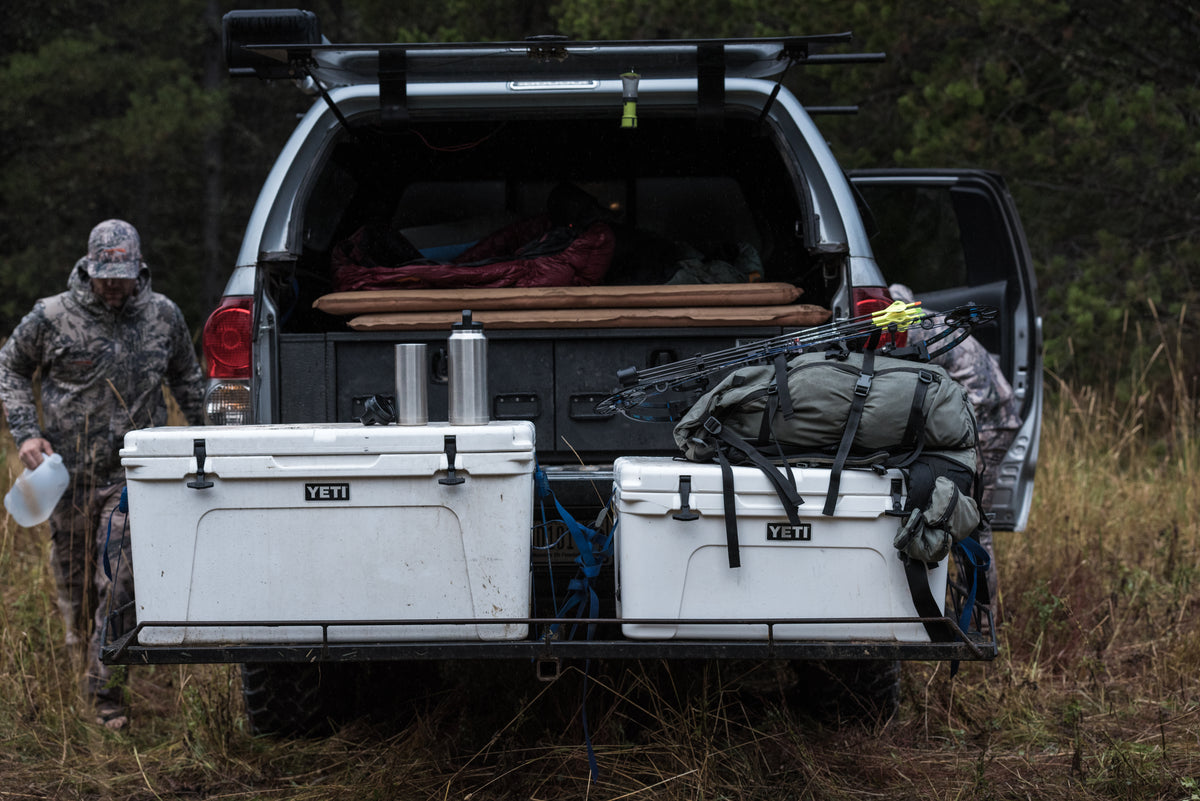 Zwei Personen in Tarnkleidung stehen neben einem offenen Geländewagen, in dessen Laderaum sich zwei große YETI®-Kühlboxen – darunter die YETI®-Kühlbox Tundra 110 in Weiß –, ein Rucksack, ein Bogen und Schlafausrüstung befinden. Die Szene spielt im Freien, umgeben von hohem Gras und Bäumen.