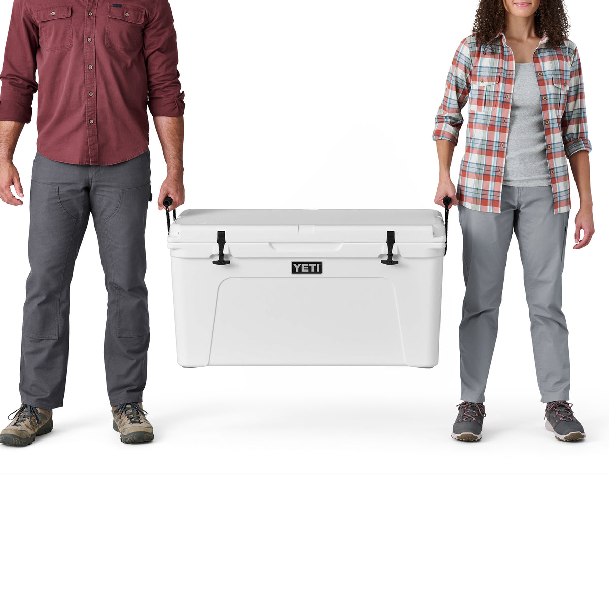 Ein Mann und eine Frau, beide in legerer Outdoor-Kleidung, stehen nebeneinander und halten eine große weiße YETI®-Kühlbox „Tundra 110“ zwischen sich, wobei jeder einen Griff festhält. Der Fokus liegt auf der YETI-Premium-Kühlbox und ihren Unterkörpern.