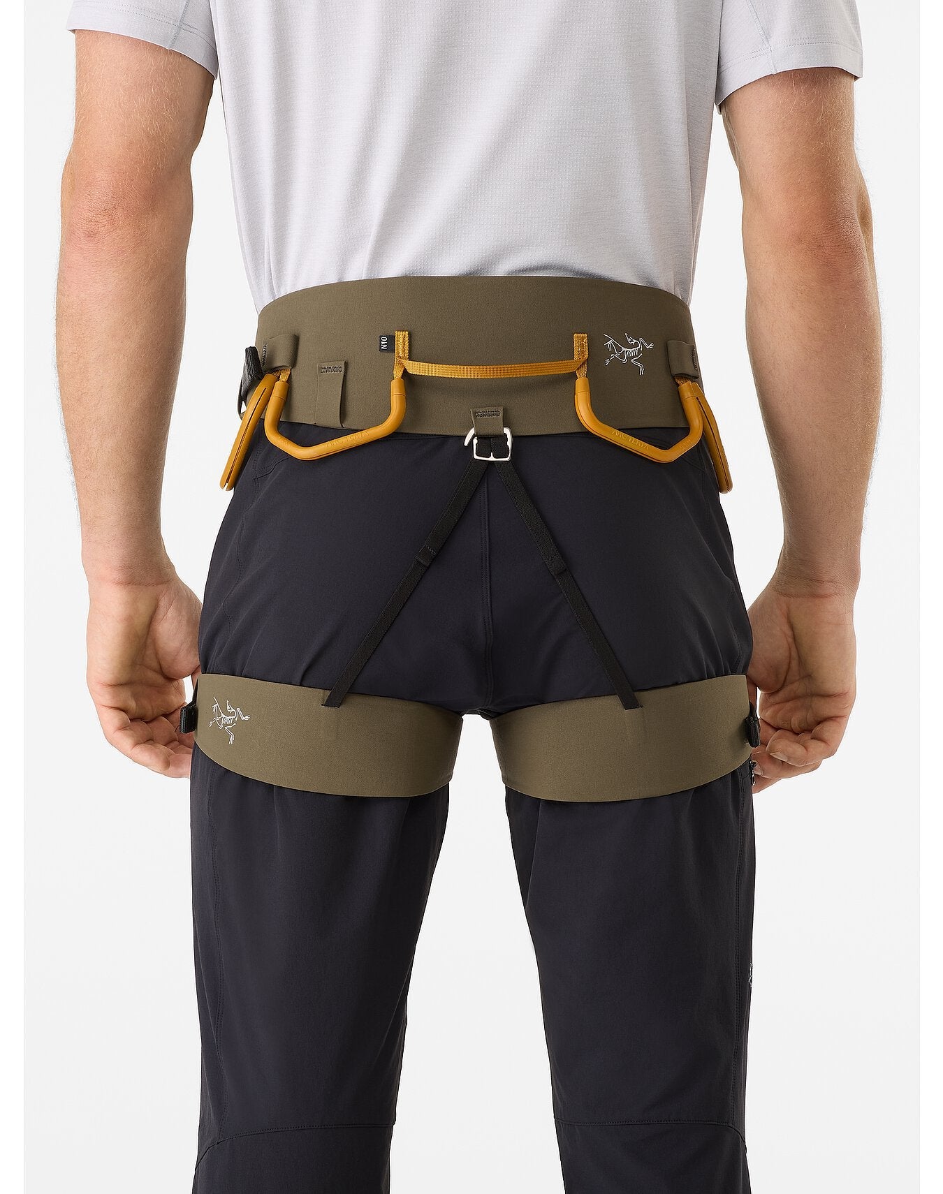 Person von hinten, die einen ARC'TERYX LEAF AR-395a Gurt in Olivgrün und Orange über einer dunklen Hose trägt. Das Gurtzeug verfügt über Materialschlaufen, Warp Strength Technology und ein sichtbares Arc'teryx-Logo. Die Hände ruhen an der Seite.