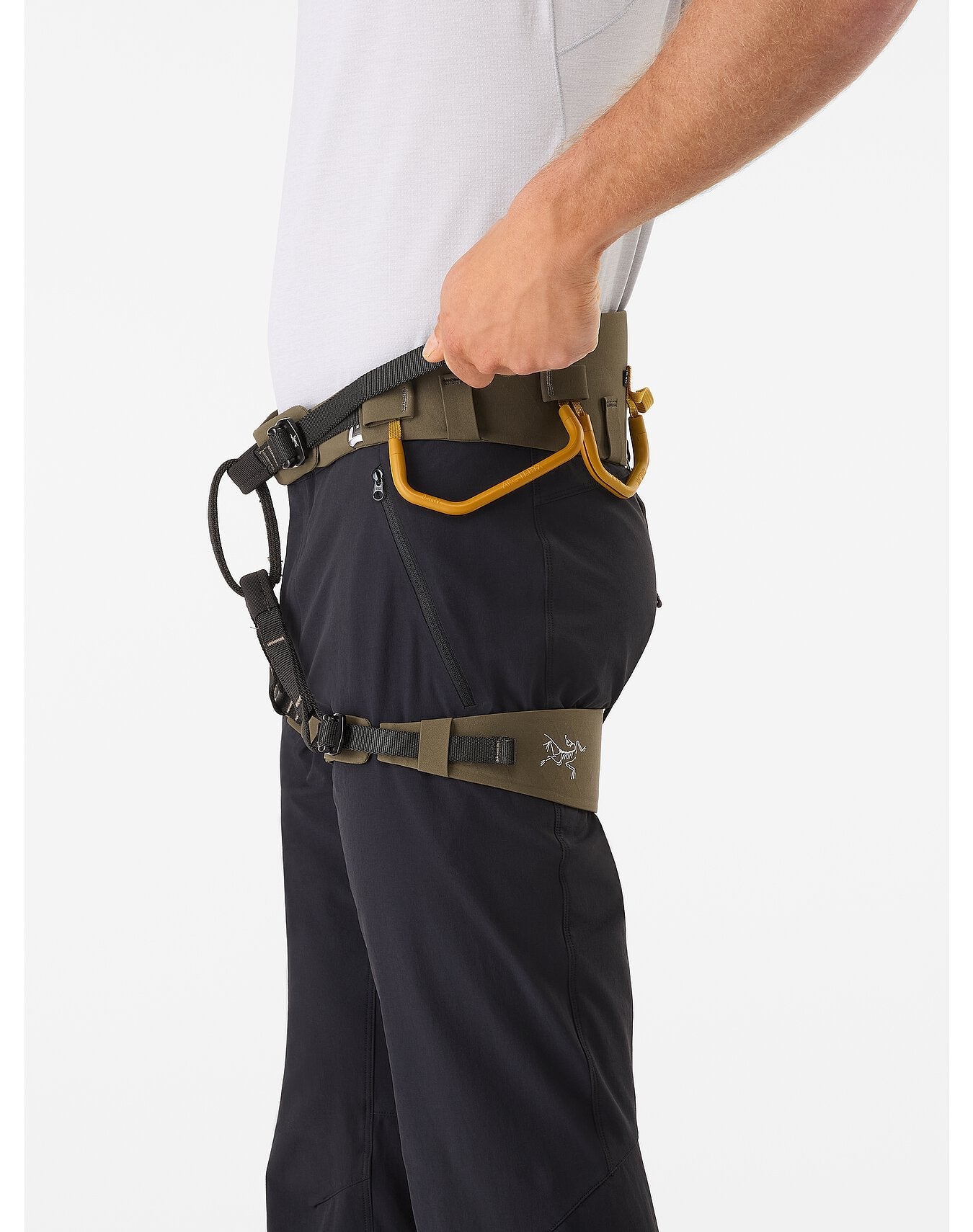 Eine Person in weißem Hemd und schwarzer Hose legt sich ein ARC'TERYX LEAF Arc'teryx AR-395a Harness with Warp Strength Technology um die Taille und die Oberschenkel und befestigt einen gelben Karabiner an dem Hochleistungsgurt für Männer.