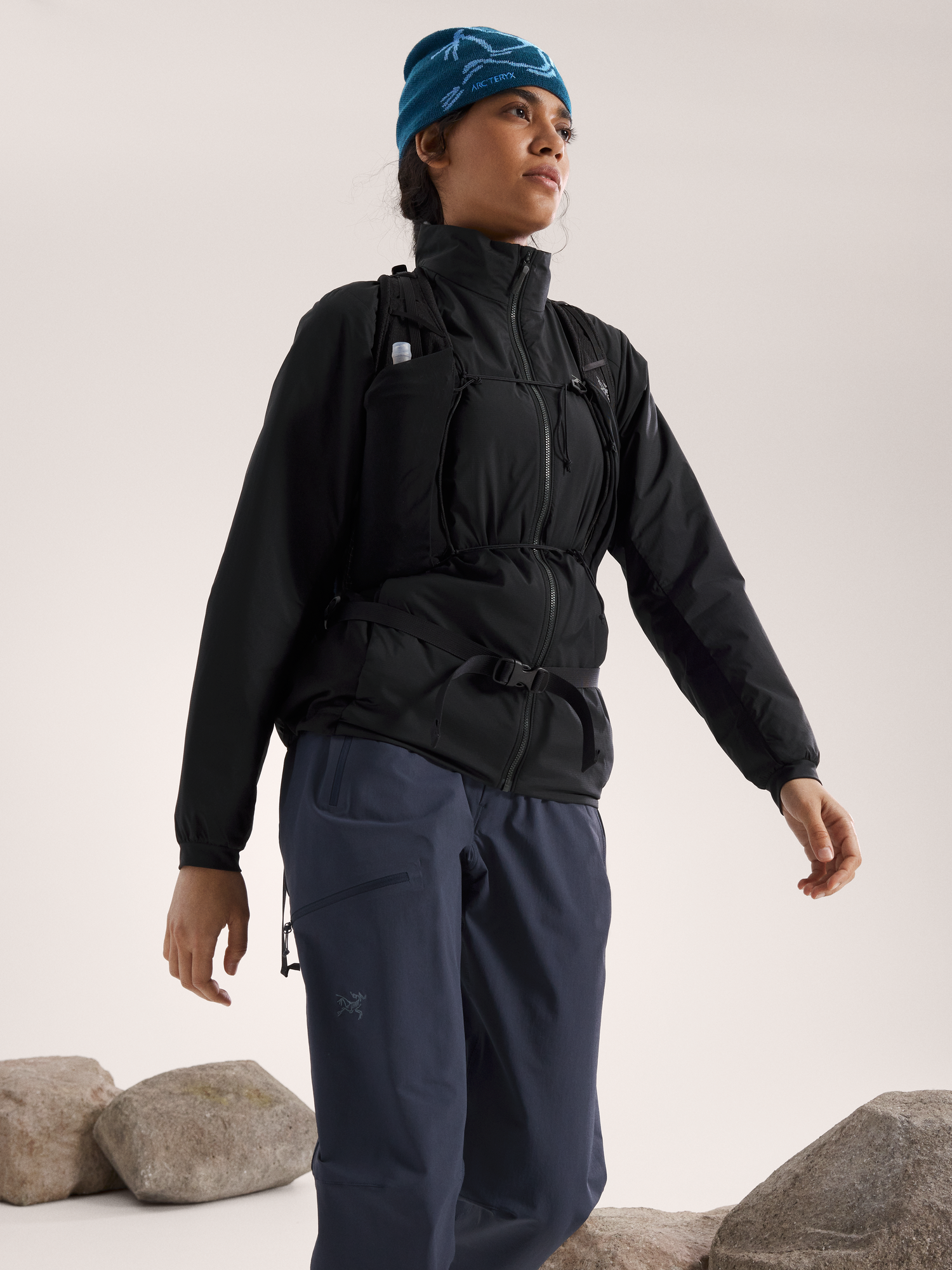 Eine Frau mit blauer Mütze, schwarzer ARC'TERYX Atom Jacket Women's - Black und blauer Outdoor-Hose steht auf Felsen vor einem neutralen Hintergrund und blickt mit ausgestrecktem Arm in die Ferne.