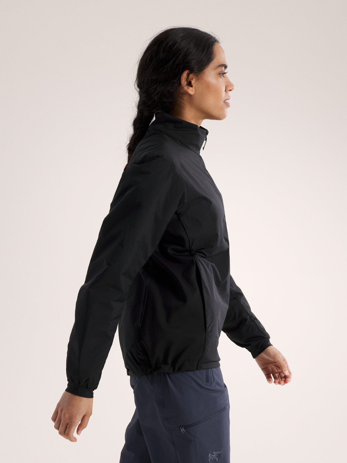 Eine Frau mit langen dunklen Haaren, die zu einem Zopf geflochten sind, trägt eine schwarze ARC'TERYX Arc'teryx Atom Jacket Women's und eine marineblaue Hose. Sie steht im Profil vor einem schlichten hellen Hintergrund.