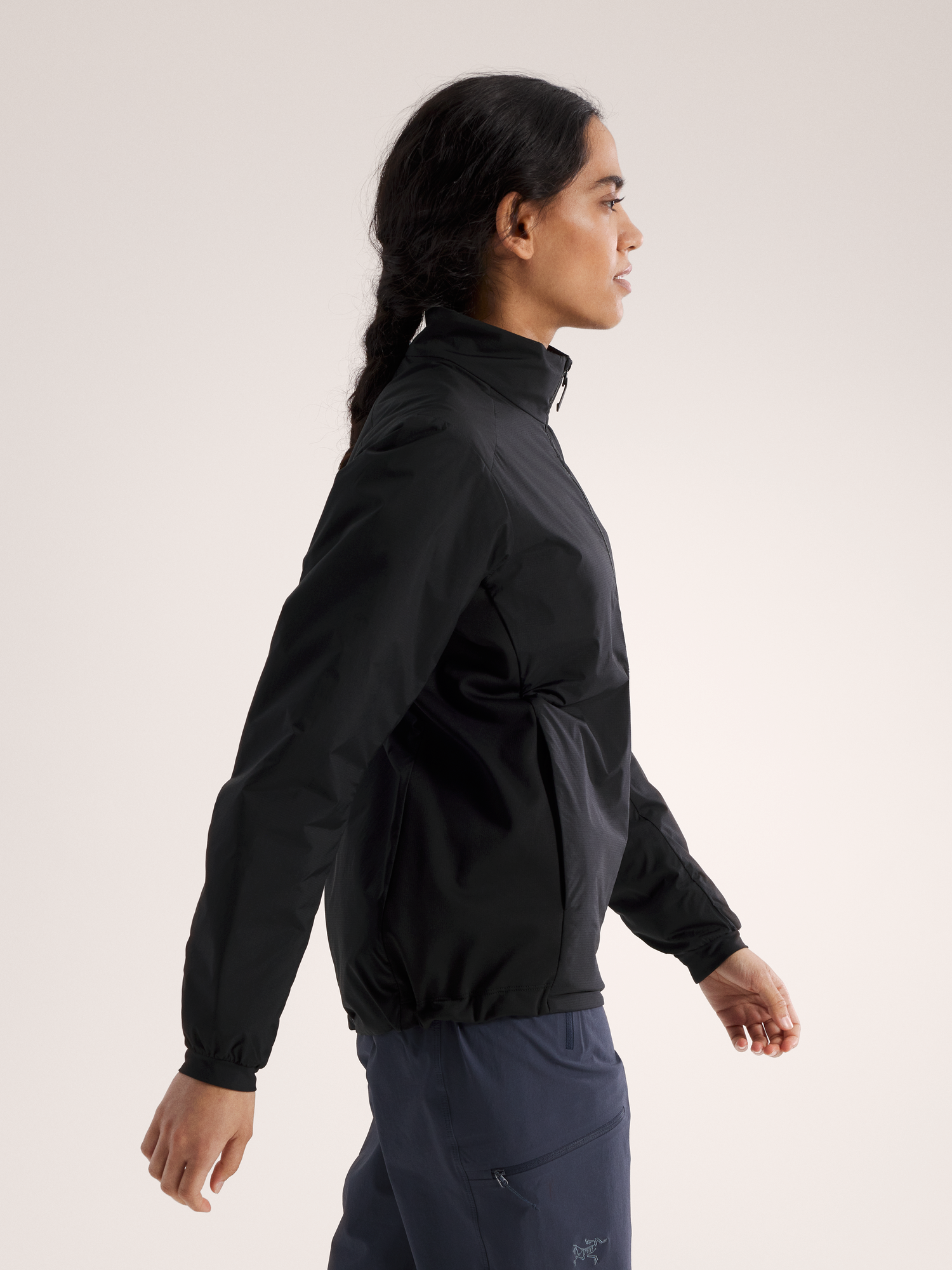Eine Frau mit langen dunklen Haaren, die zu einem Zopf geflochten sind, trägt eine schwarze ARC'TERYX Arc'teryx Atom Jacket Women's und eine marineblaue Hose. Sie steht im Profil vor einem schlichten hellen Hintergrund.