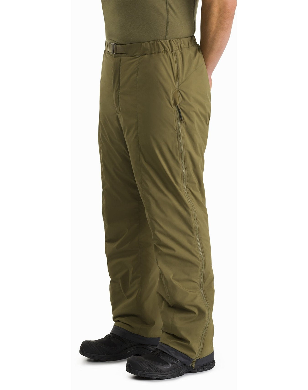 Eine Person, die ARC'TERYX LEAF Arc'teryx Atom Pant LT Gen2 in Crocodile mit einem passenden grünen Hemd und schwarzen Schuhen trägt, steht mit den Händen in den Taschen vor einem weißen Hintergrund.