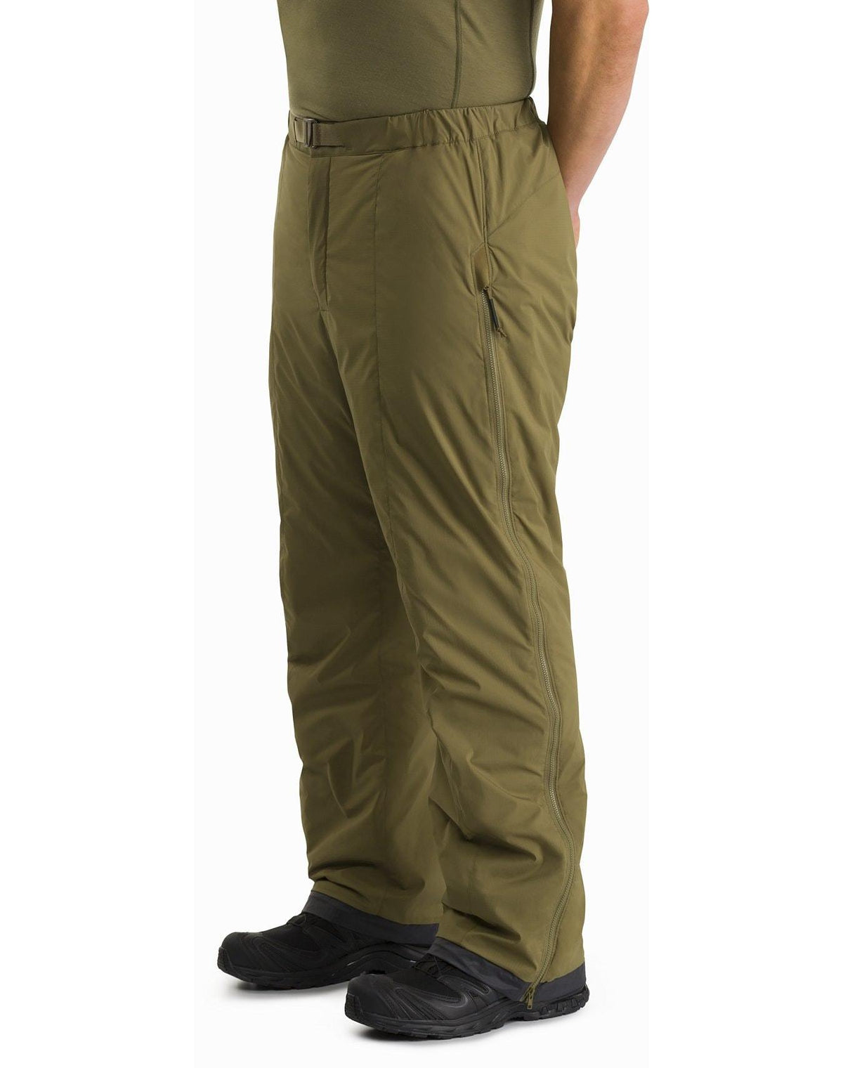 Eine Person, die die schwarze ARC'TERYX LEAF Arc'teryx Atom Pant LT Gen2, ein grünes Hemd und schwarze Schuhe trägt, steht mit den Händen in den Taschen. Die locker sitzende Hose ist ideal für den Einsatz im Freien oder bei kaltem Wetter.