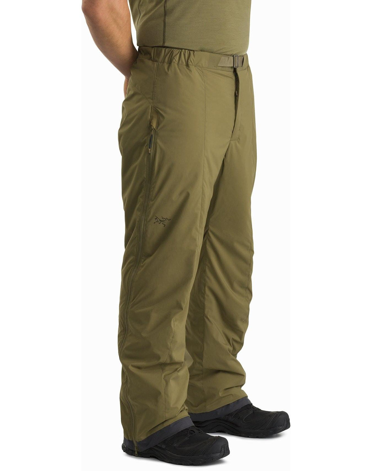 Eine Person trägt eine schwarze ARC'TERYX LEAF Arc'teryx Atom Pant LT Gen2, eine leichte Outdoor-Hose mit Reißverschlusstaschen, gepaart mit einem grünen Hemd und schwarzen Wanderschuhen und steht mit den Händen in den Taschen.