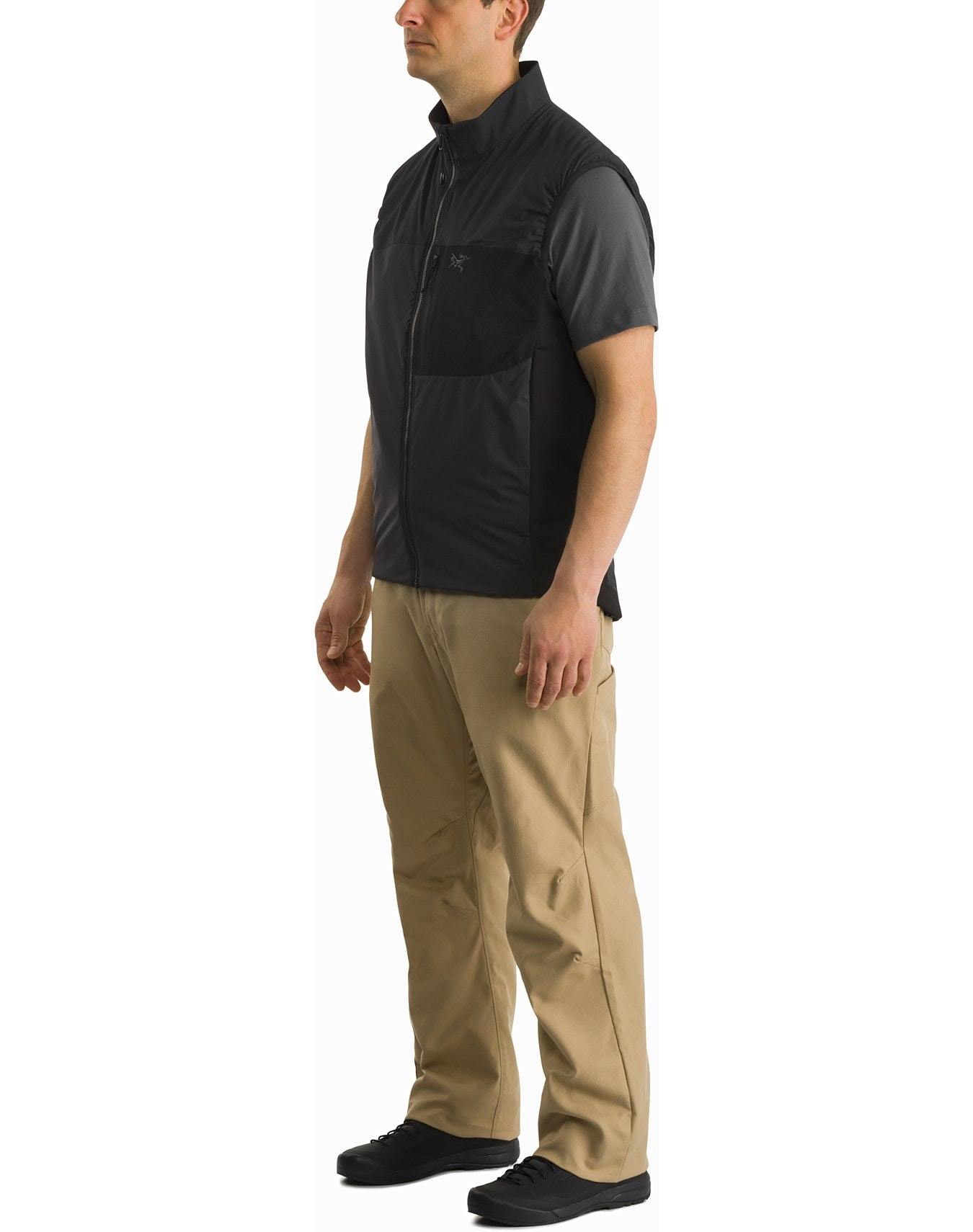 Ein Mann steht leicht gedreht, trägt ein kurzärmeliges graues Hemd, ARC'TERYX LEAF Arc'teryx Atom Vest LT Gen2.1 in Schwarz, eine beigefarbene Hose und schwarze Schuhe vor einem weißen Hintergrund.