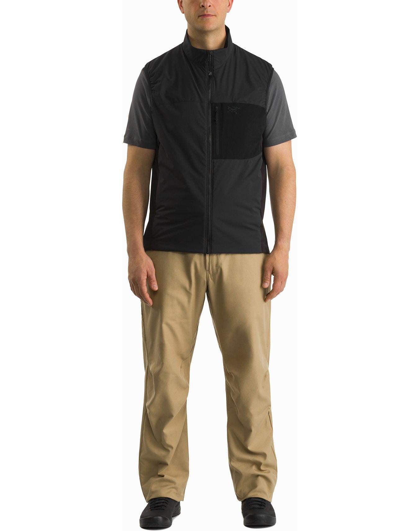 Ein Mann steht mit dem Gesicht nach vorne und trägt eine ARC'TERYX LEAF Arc'teryx Atom Vest LT Gen2.1 in Schwarz über einem grauen Hemd, mit einer hellbraunen Hose und schwarzen Schuhen, vor einem schlichten weißen Hintergrund.