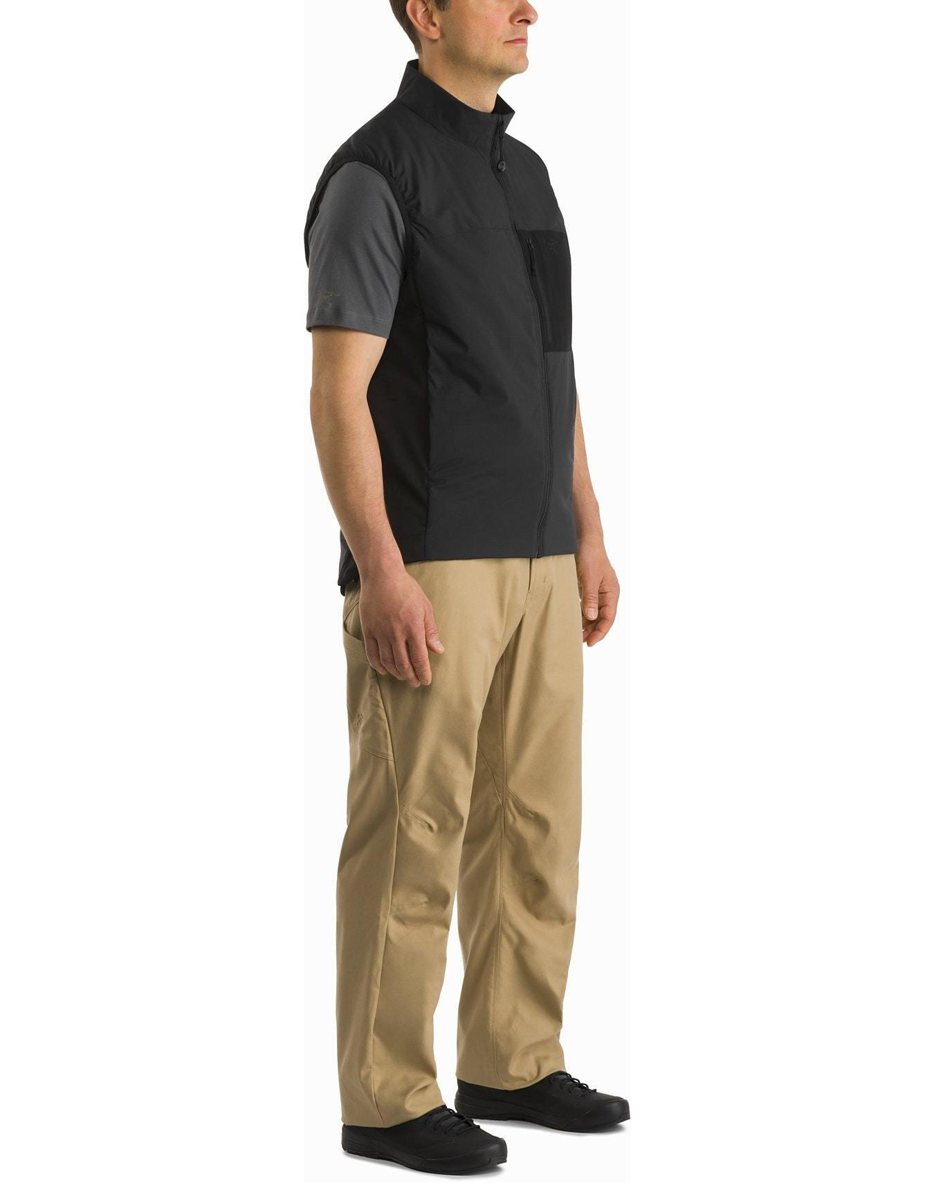 Ein Mann steht leicht nach links vor einem weißen Hintergrund. Er trägt ein kurzärmeliges graues Hemd, ARC'TERYX LEAF Arc'teryx Atom Vest LT Gen2.1 - Black, eine beigefarbene Hose und schwarze Schuhe.