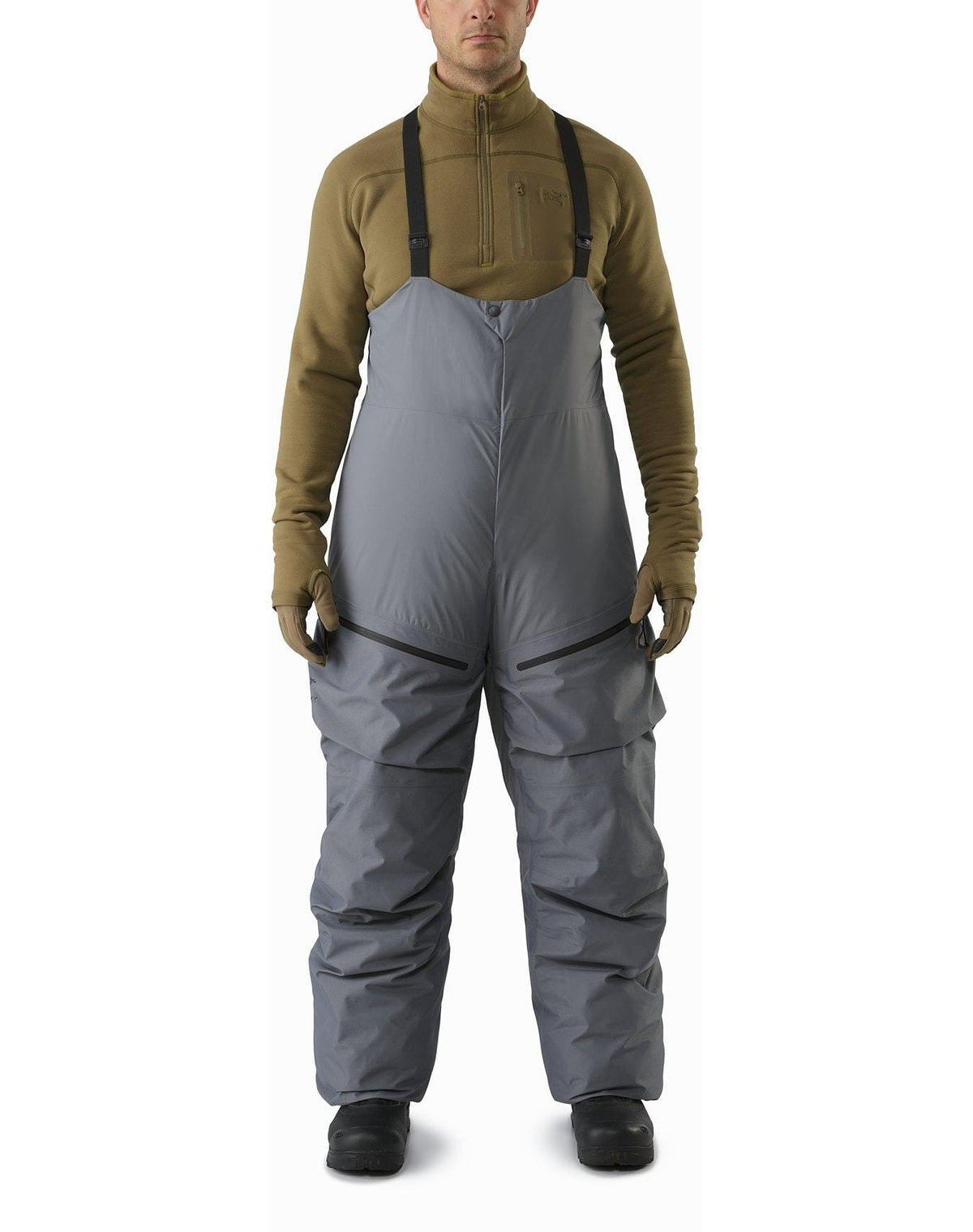 Eine Person, die einen hellbraunen Fleecepullover, hellbraune Handschuhe, ARC'TERYX LEAF Arc'teryx Cold WX Bib Pant SVX Men's - Harrier mit schwarzen Hosenträgern und schwarze Stiefel trägt, steht vor einem weißen Hintergrund.