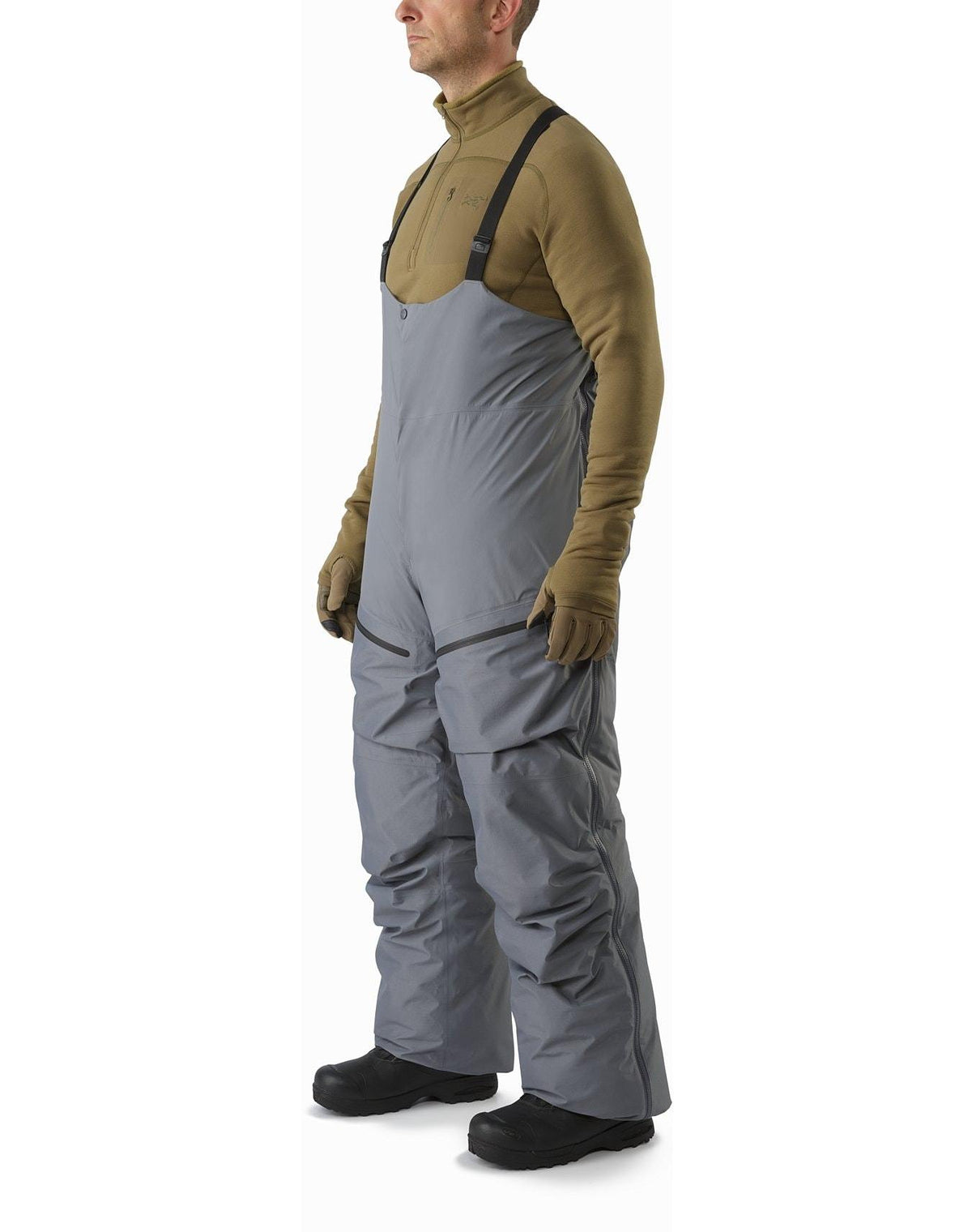 Mann trägt ARC'TERYX LEAF Arc'teryx Cold WX Bib Pant SVX Men's - Harrier (grau), mit schwarzen Hosenträgern, olivgrünem Fleecepullover, braunen Handschuhen und schwarzen Schneestiefeln. Seitenansicht auf weißem Hintergrund.