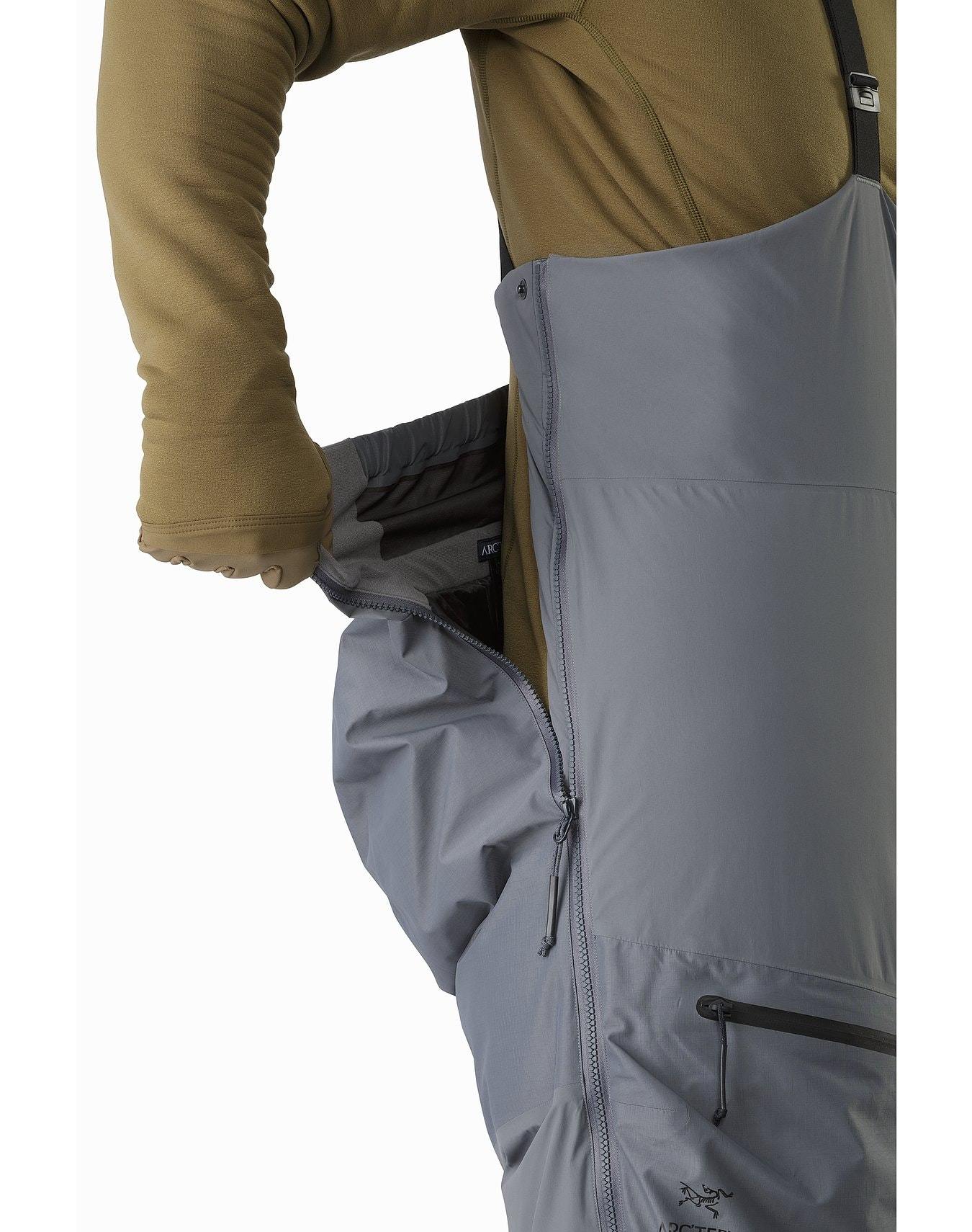 Eine Person in einem braunen, langärmeligen Hemd und einer grauen ARC'TERYX LEAF Arc'teryx Cold WX Bib Pant SVX Men's - Harrier öffnet einen seitlichen Reißverschluss an der Hose, um eine innere Schicht freizulegen. Der Hintergrund ist einfarbig weiß.