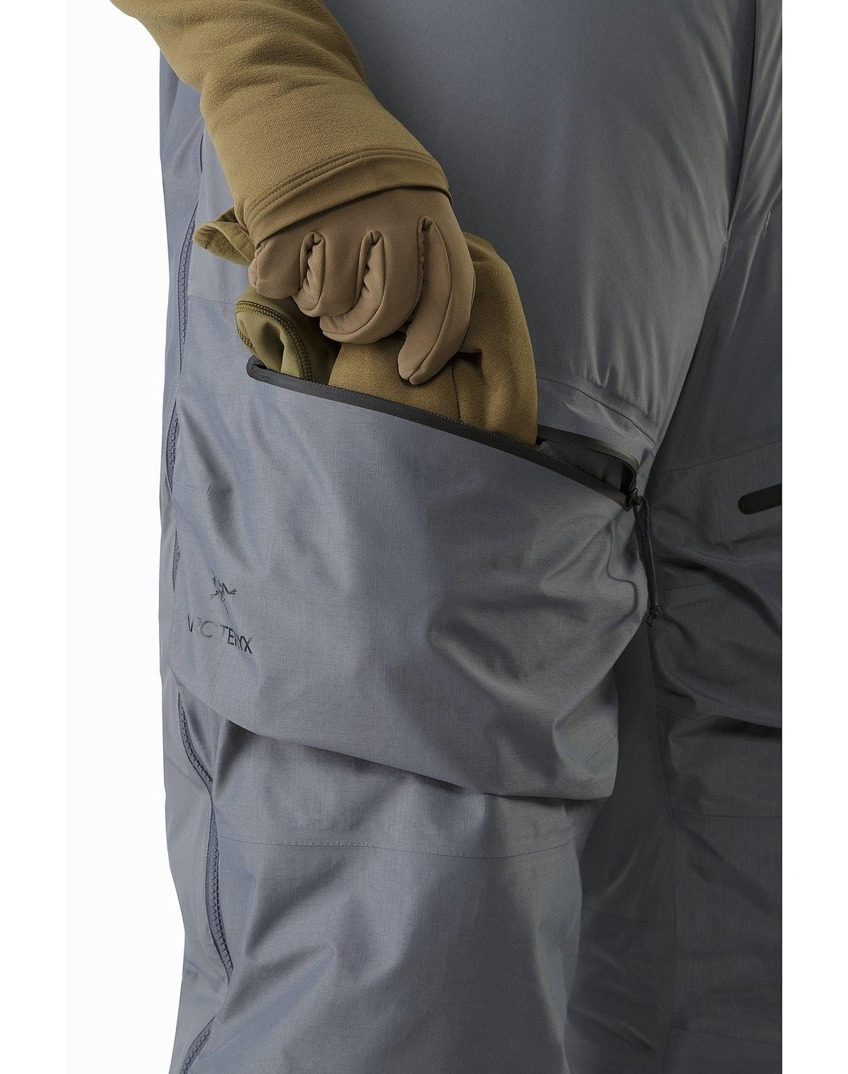 Eine Person mit hellbraunen Handschuhen und grauer Jacke steckt ihre behandschuhte Hand in die Seitentasche mit Reißverschluss der grauen Arc'teryx Cold WX Bib Pant SVX Men's - Harrier von ARC'TERYX LEAF, die für Komfort und Wärme bei kalten Bedingungen entwickelt wurde.
