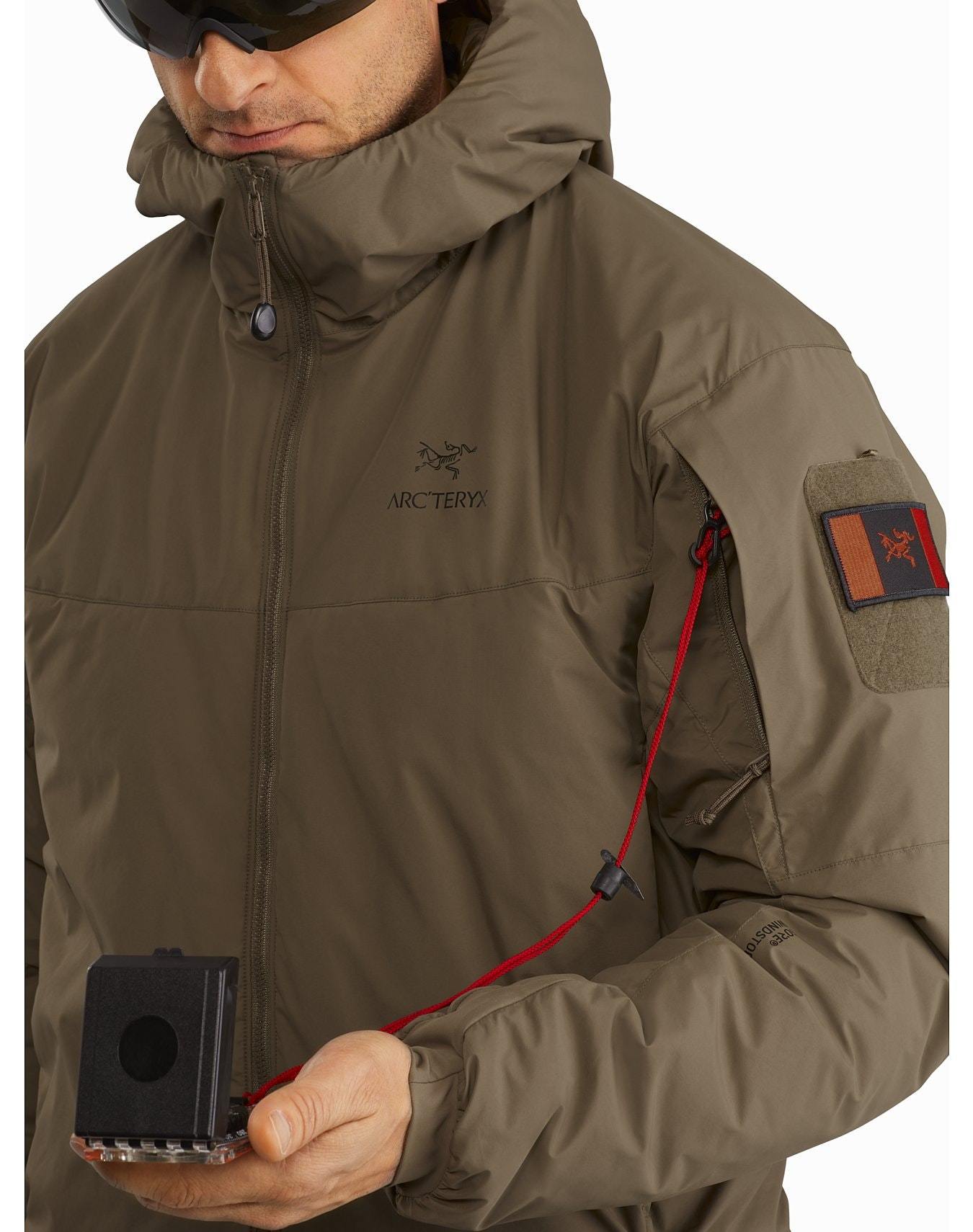 Eine Person, die ein ARC'TERYX LEAF Cold WX Hoody LT Gen2.1 - Crocodile und eine Sonnenbrille trägt, hält ein kleines schwarzes Gerät, das mit einer roten Schnur an der oberen Ärmeltasche der Jacke befestigt ist. Arc'teryx-Logos sind auf der Brust und dem Ärmelaufnäher zu sehen.