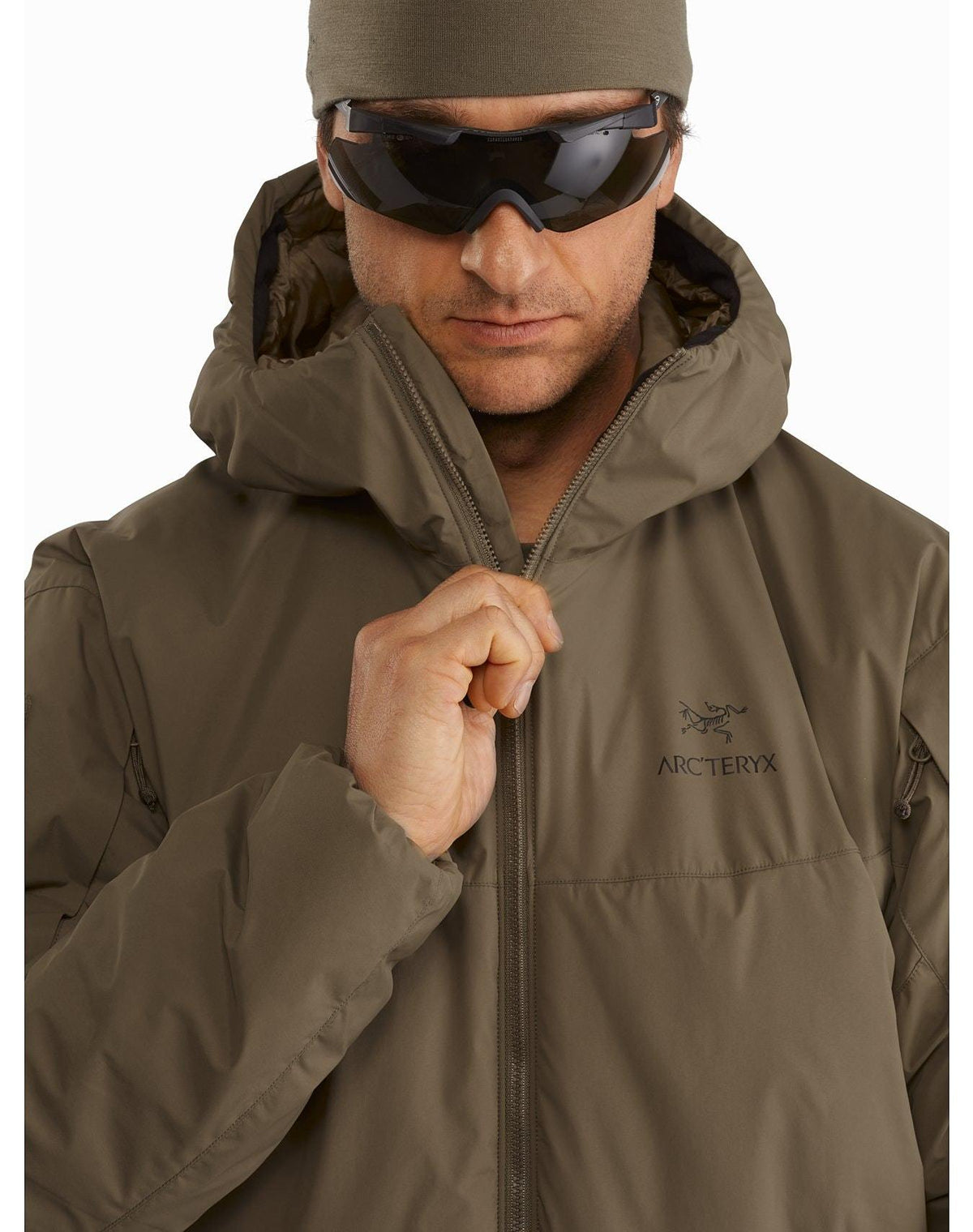 Eine Person, die einen rangergrünen ARC'TERYX LEAF Cold WX Hoody LT Gen2.1, eine dunkle Sonnenbrille und eine braune Mütze trägt, schließt mit neutraler Miene den Reißverschluss der Jacke.