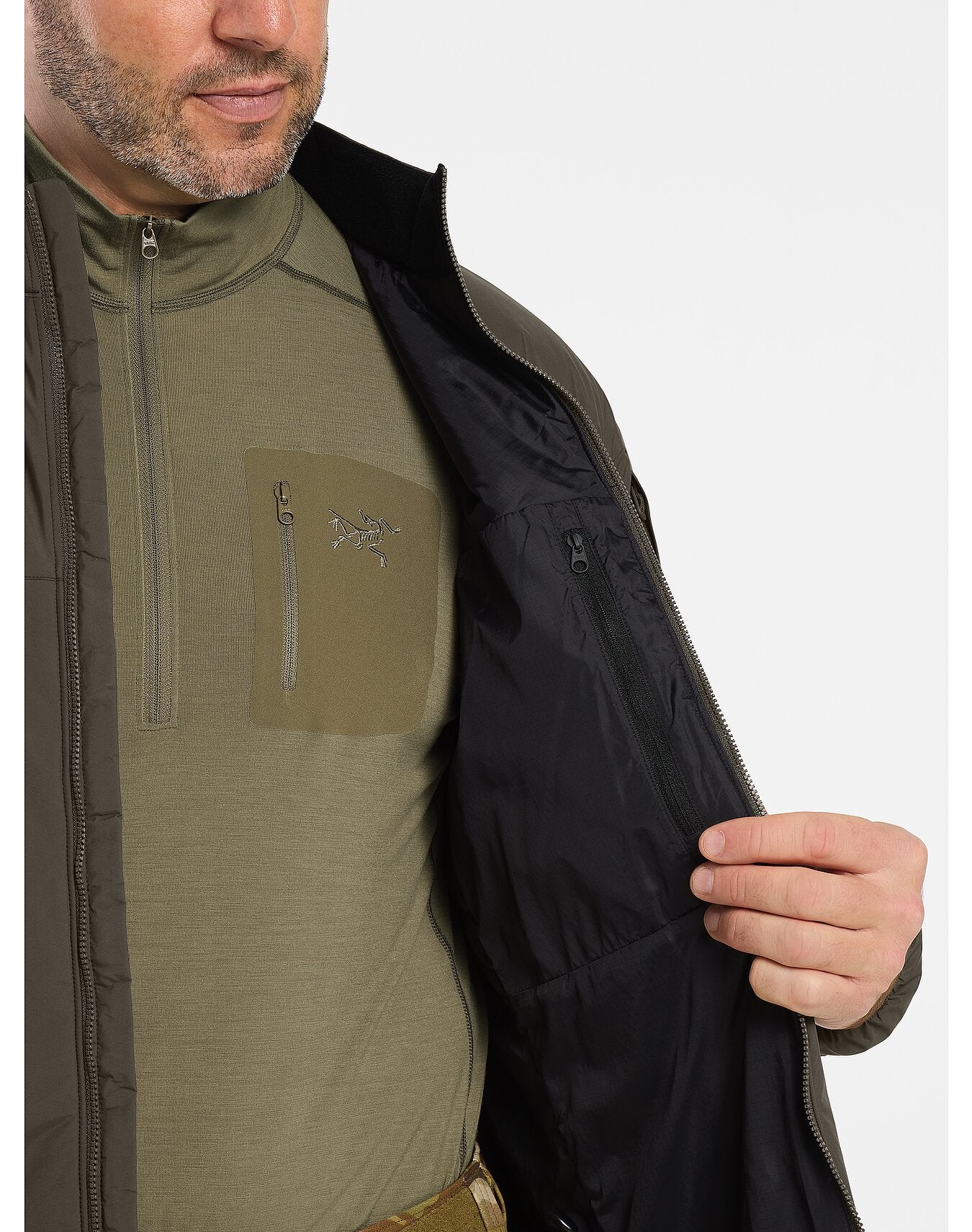 Ein Mann öffnet den Reißverschluss seiner schwarzen Arc'teryx Cold WX Jacket LT Gen2.1 by ARC'TERYX LEAF. Zum Vorschein kommt ein grünes Reißverschlusshemd mit Brusttasche und weißem Logo. Er zeigt das schwarze Futter der Jacke, die Innentasche mit Reißverschluss und den GORE WINDSTOPPER Schutz.