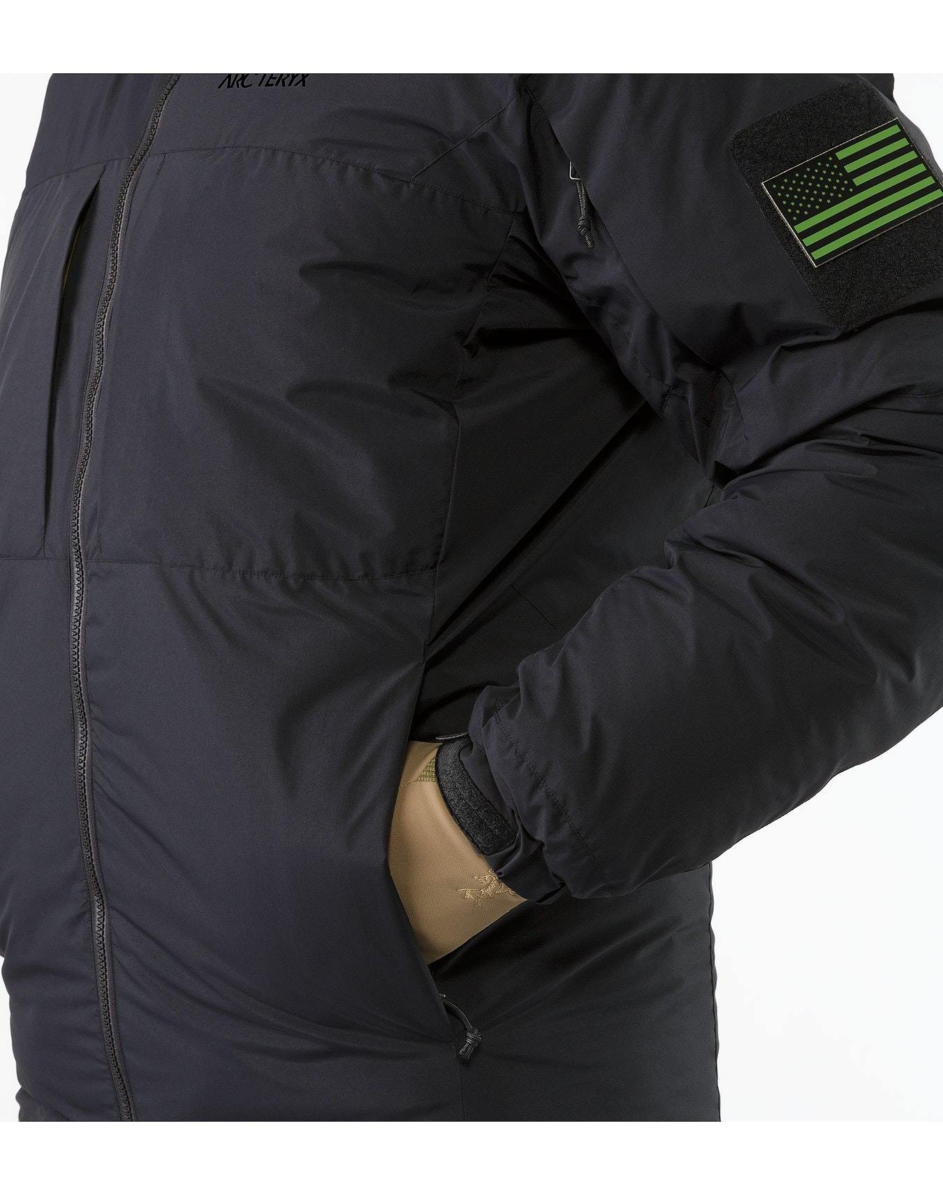 Eine Person, die vom Oberkörper bis zum rechten Arm vor weißem Hintergrund zu sehen ist, trägt eine ARC'TERYX LEAF Arc'teryx Cold WX Jacket SV Gen1.4 mit einem grün-schwarzen Aufnäher der amerikanischen Flagge auf dem Ärmel und der Hand in der Tasche.